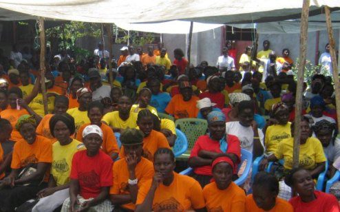 A large group of people seated under a canopy, wearing colorful shirts in red, orange, yellow, and blue, suggesting a communal or event gathering.