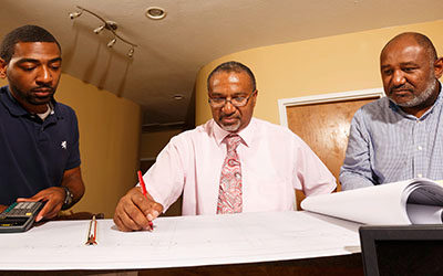 Three people reviewing architectural plans on a table, with one person writing with a pen. They appear to be discussing or planning a project.