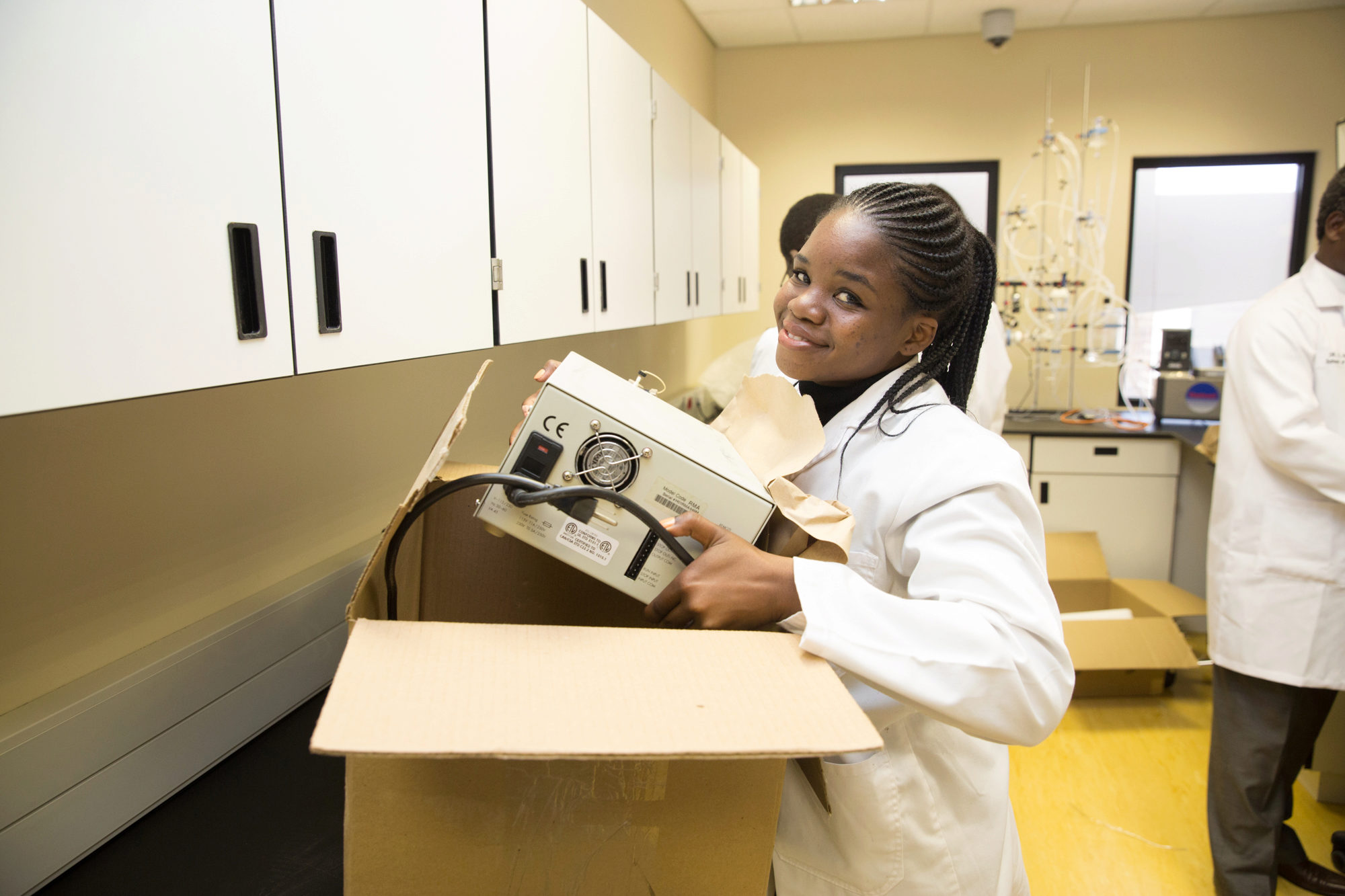 A person in a lab coat holding laboratory equipment, smiling while unpacking a box in a laboratory setting with cabinets and lab apparatus in the background.