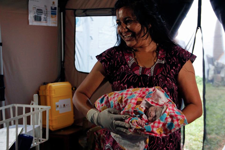 A person smiling and holding a newborn wrapped in a colorful blanket inside a tent-like setting, suggesting a field hospital or medical camp.