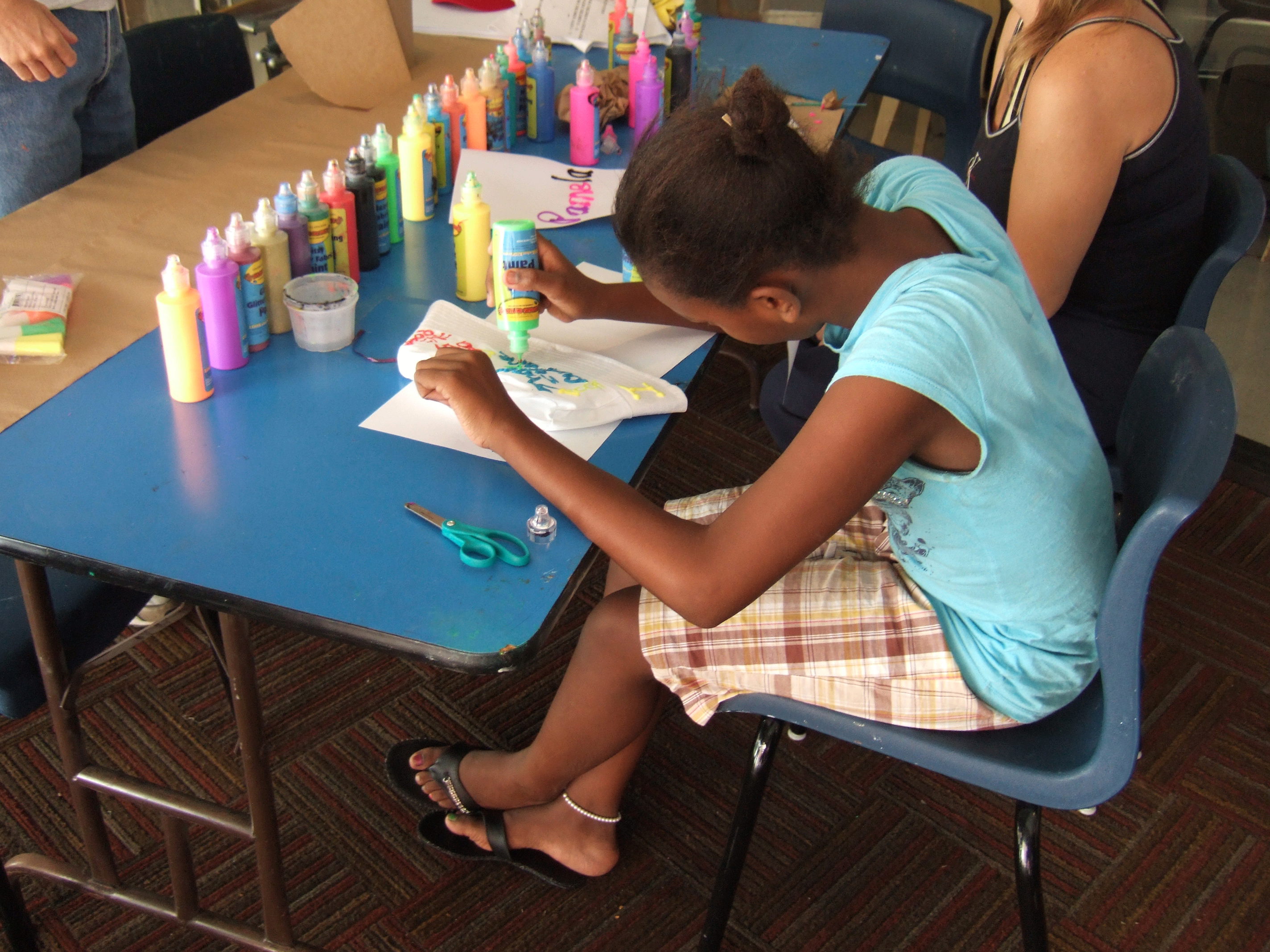 A child seated at a table, engaging in a painting activity with various colorful paint bottles and supplies spread out.