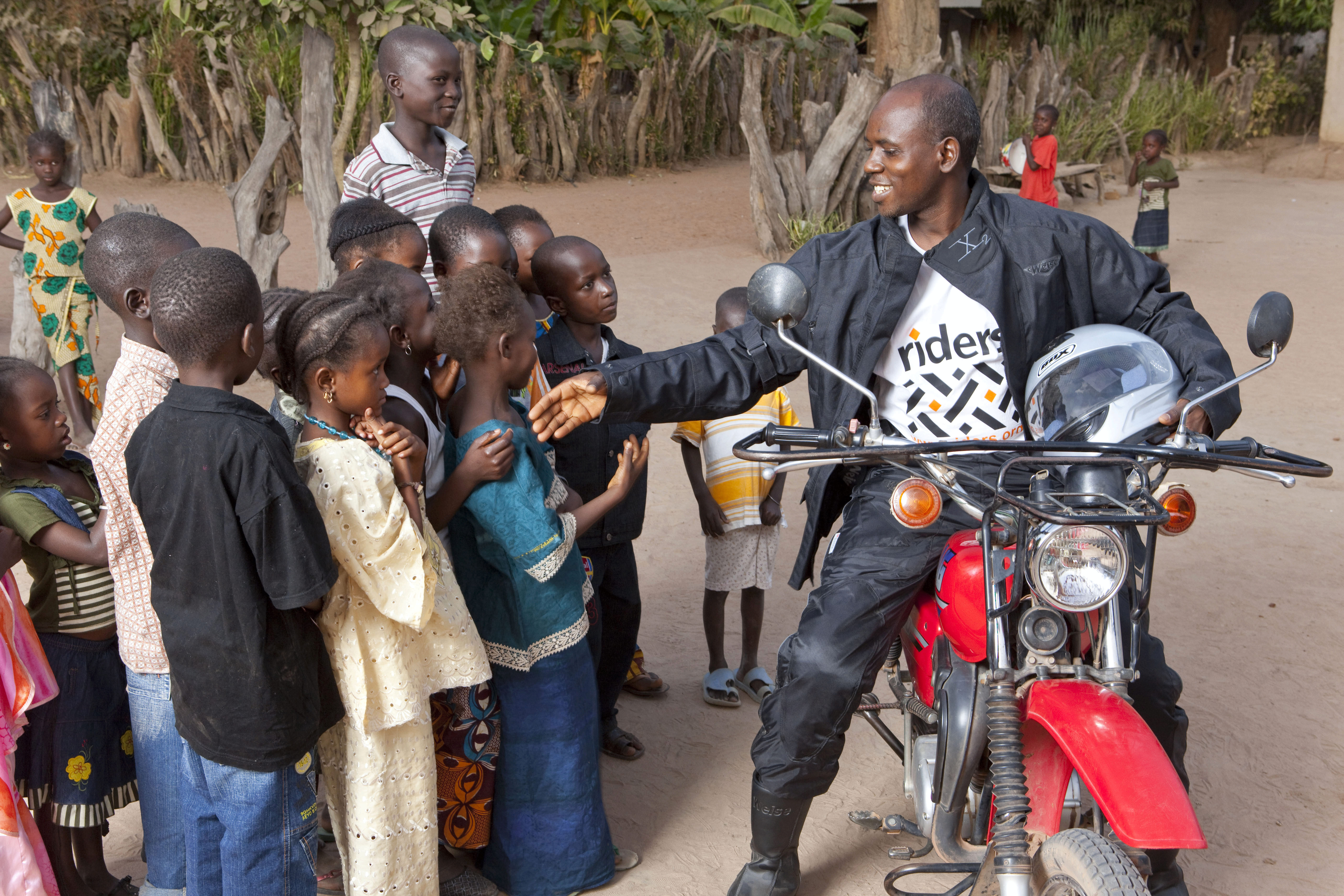 A person on a motorcycle is smiling and interacting with a group of children gathered around. The scene suggests a sense of community and positive engagement.