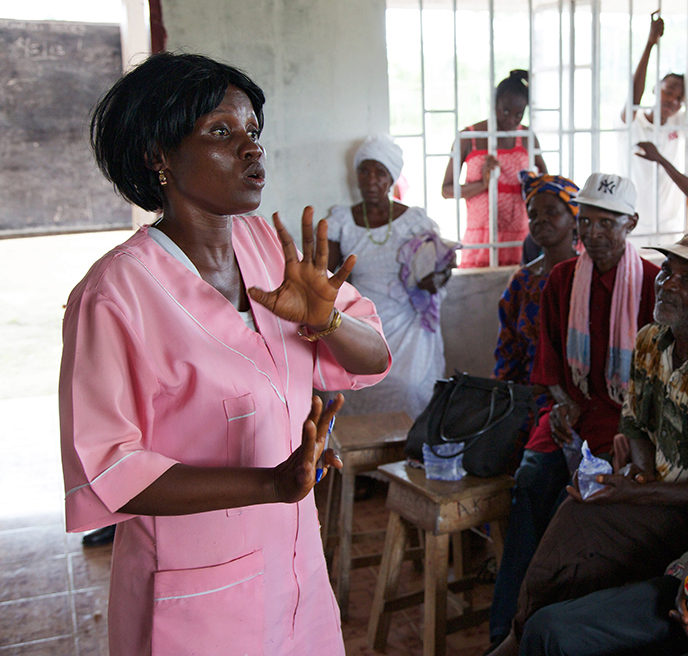 A healthcare worker in a pink uniform is speaking to a group of seated individuals in a room. The setting appears to be a community or educational session.