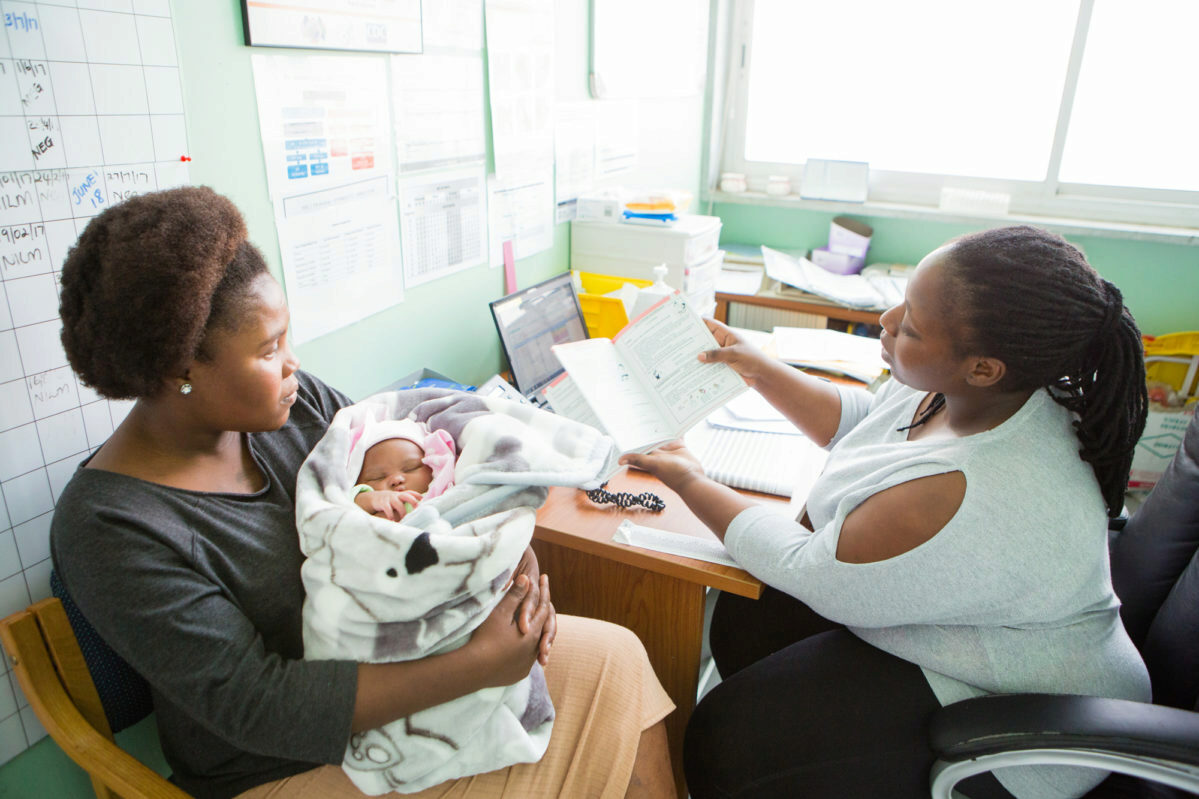 A person holding a baby wrapped in a blanket sits across from another person who is showing them a document in an office-like setting. This scene represents a consultation or meeting, possibly in a healthcare or social service environment.