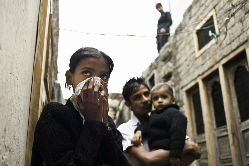 A person covering their nose with a cloth while another holds a child, standing in an alley between stone buildings. A third person is on a rooftop in the background.