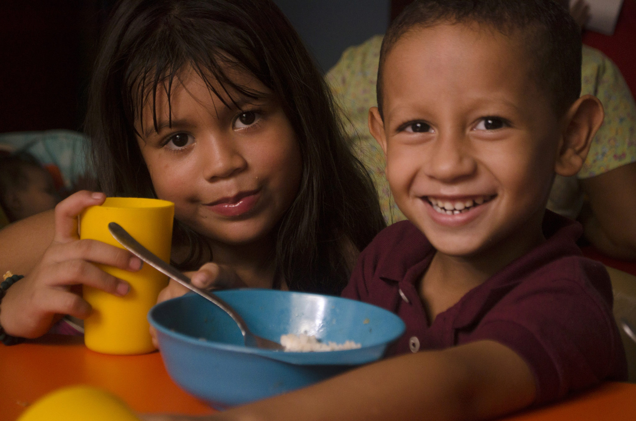 A smiling young child and another child with long hair are sitting together, holding a yellow cup and a blue bowl with a spoon. They appear to be enjoying a meal.