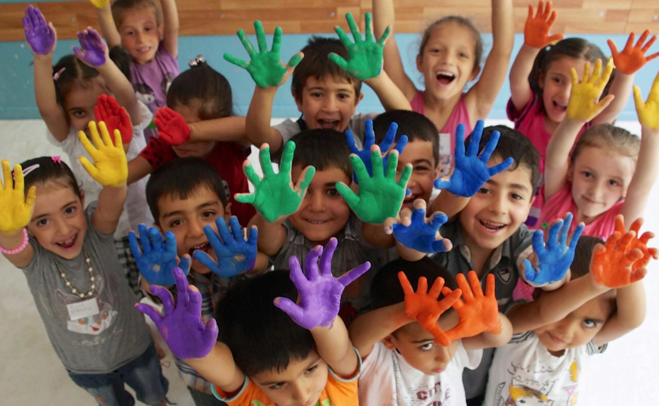 A joyful group of children with brightly painted hands in various colors, raising their hands and smiling at the camera, suggesting a fun and creative activity.