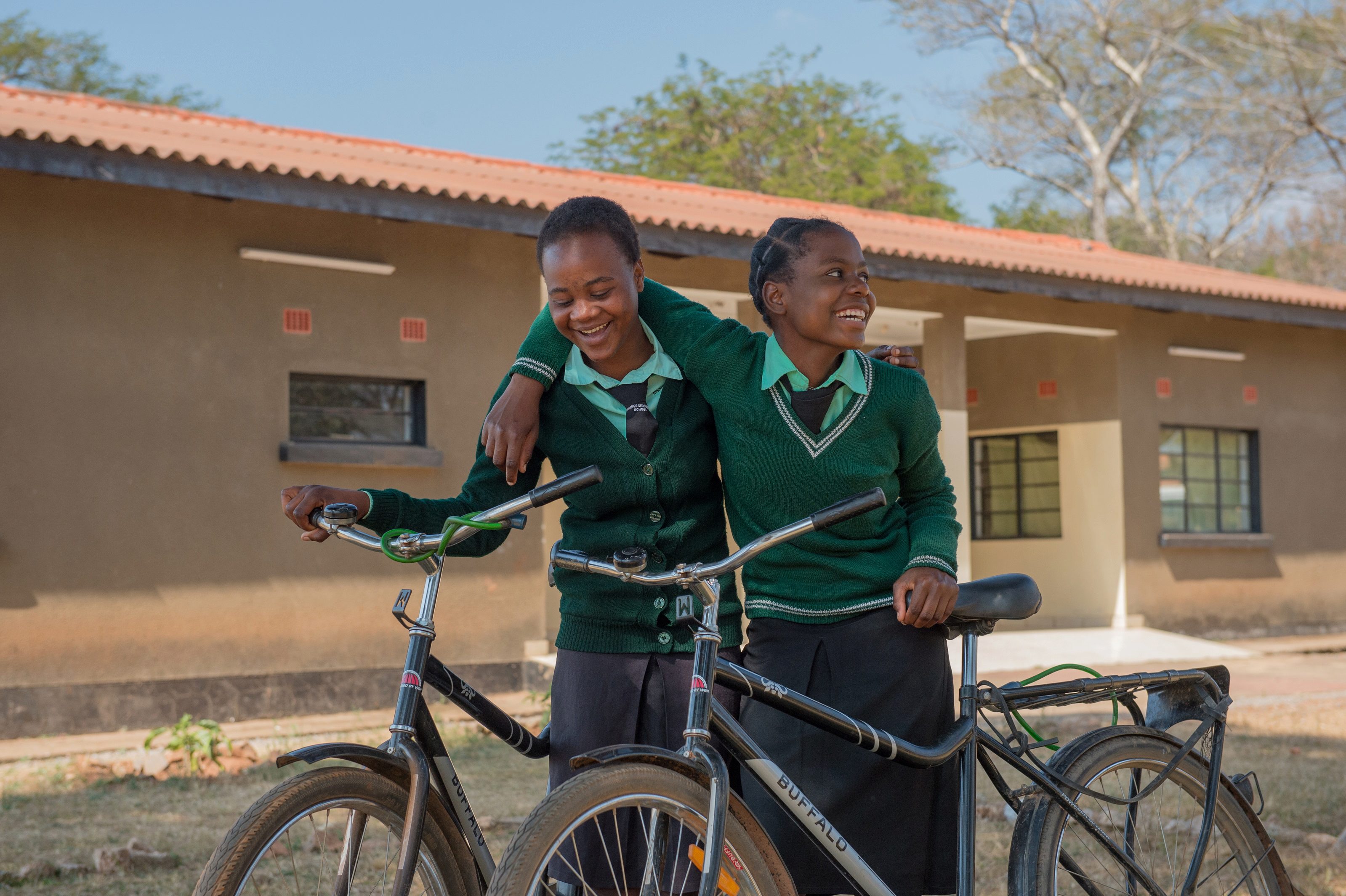 Two students in school uniforms, smiling and standing with bicycles, with one arm around each other in front of a school building.