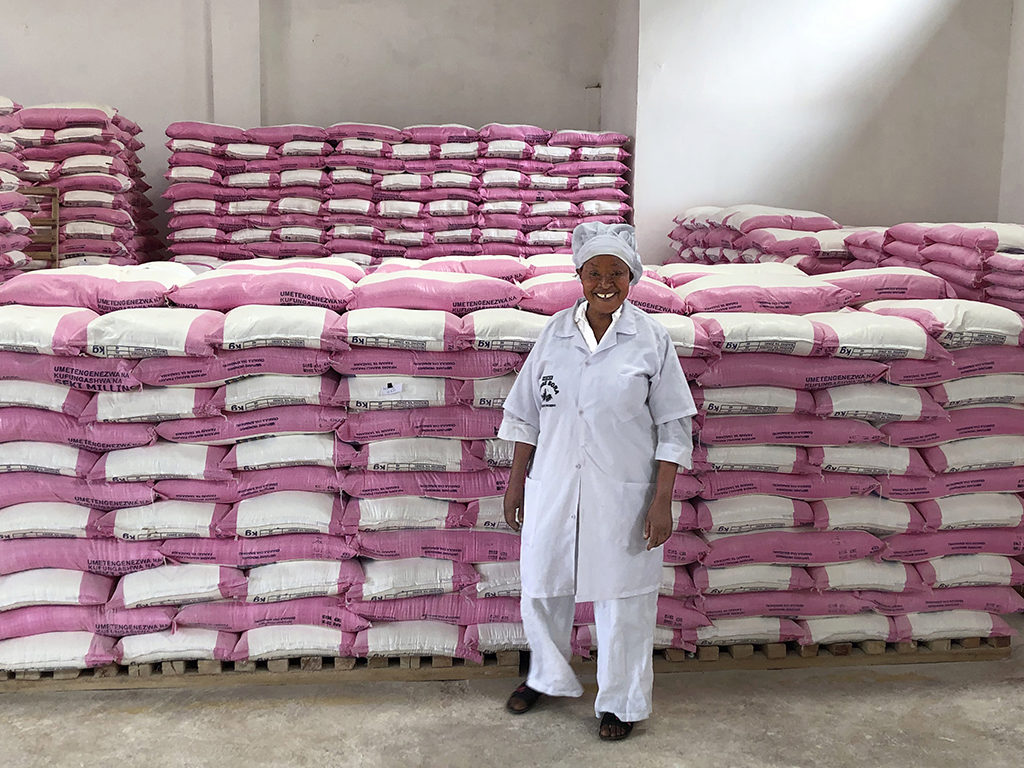 A person in a white coat and cap stands smiling in front of stacked bags of flour with pink and white packaging, inside a warehouse.