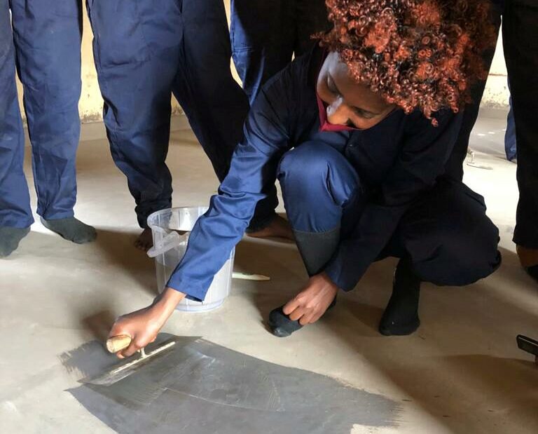 A person in a blue jumpsuit is kneeling on a floor, applying a gray substance with a trowel. A bucket is nearby, and others in similar attire are around them.