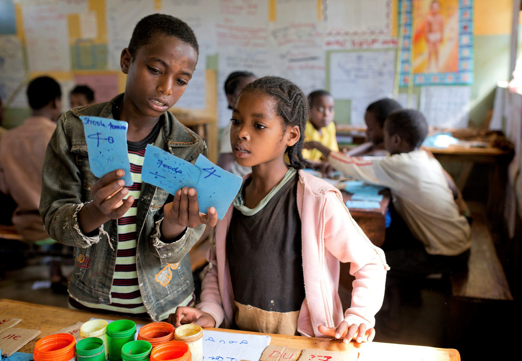 Children in a classroom are examining blue cards with writing, surrounded by colorful jars. Other students are engaged in group activities at tables in the background.