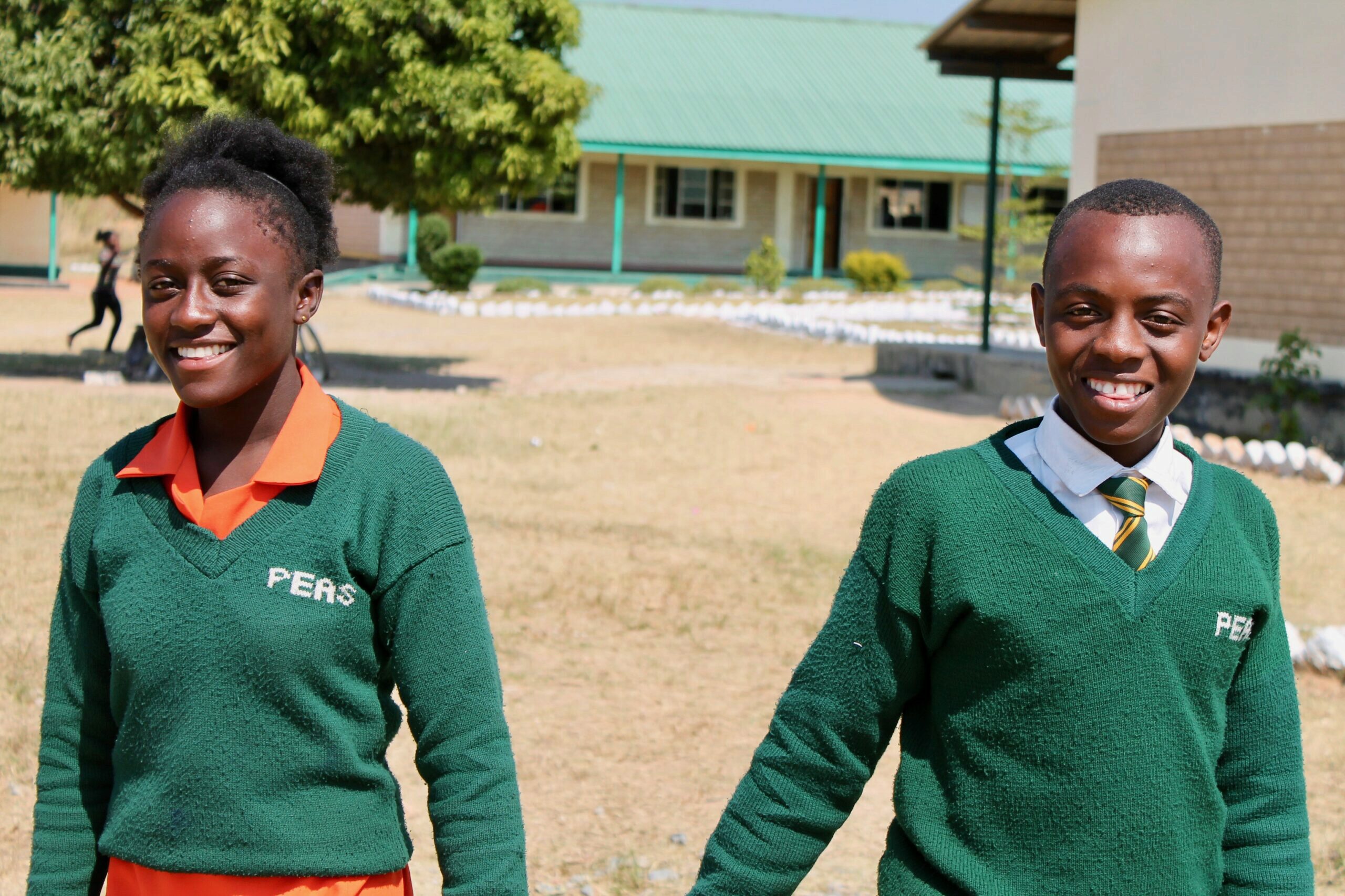 Two students, wearing school uniforms with green sweaters and logos, smiling and standing outdoors in a school courtyard.