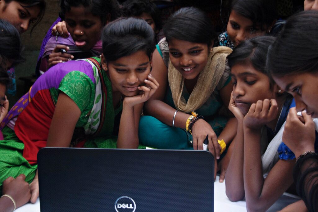 A small group of young people are gathered around a laptop, appearing engaged and focused, likely sharing or learning something together.