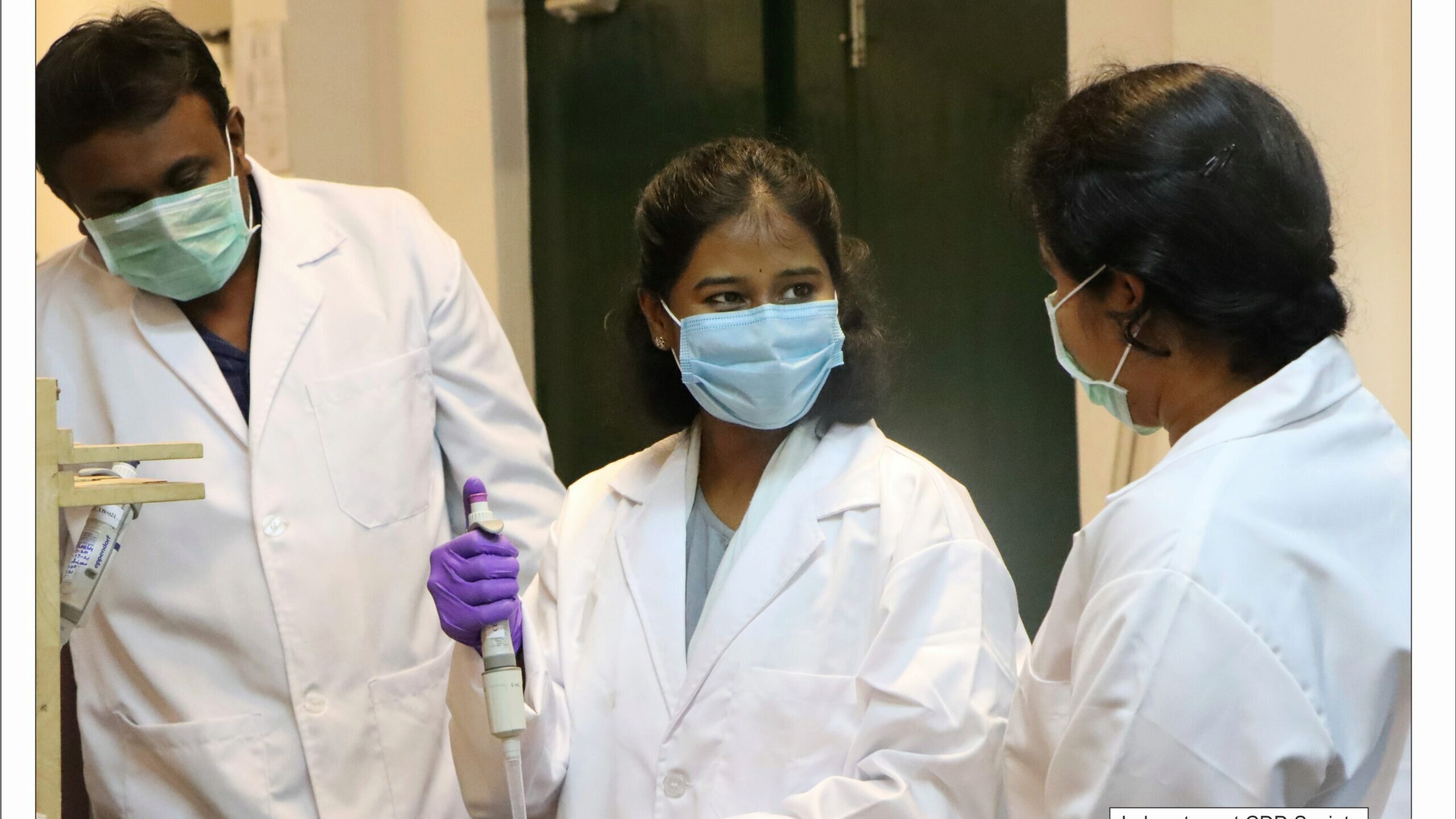 A small group of people wearing lab coats and masks are engaged in a discussion in a laboratory. One person is holding a pipette.