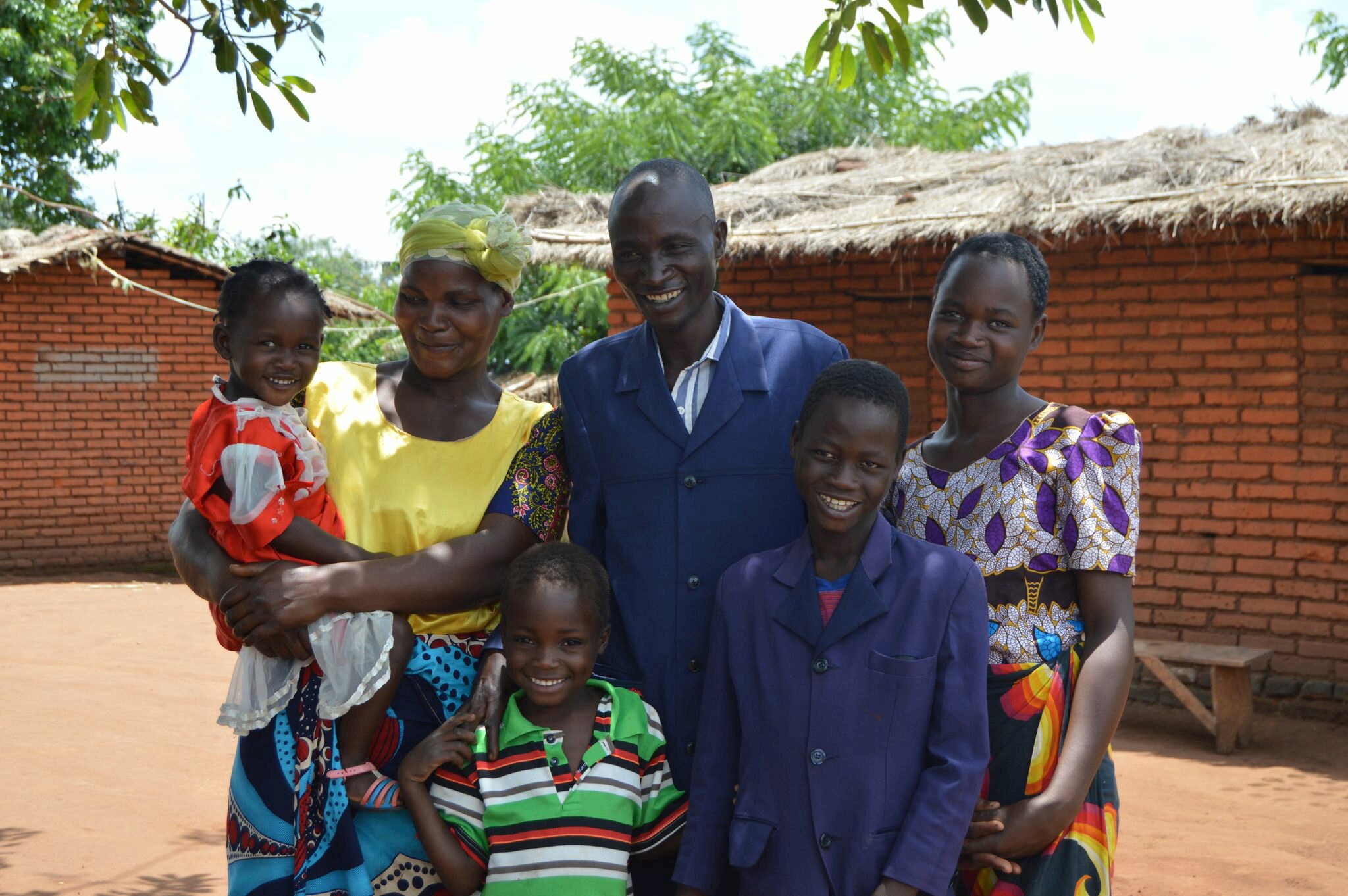 A smiling family group stands together outdoors, with brick buildings and trees in the background.