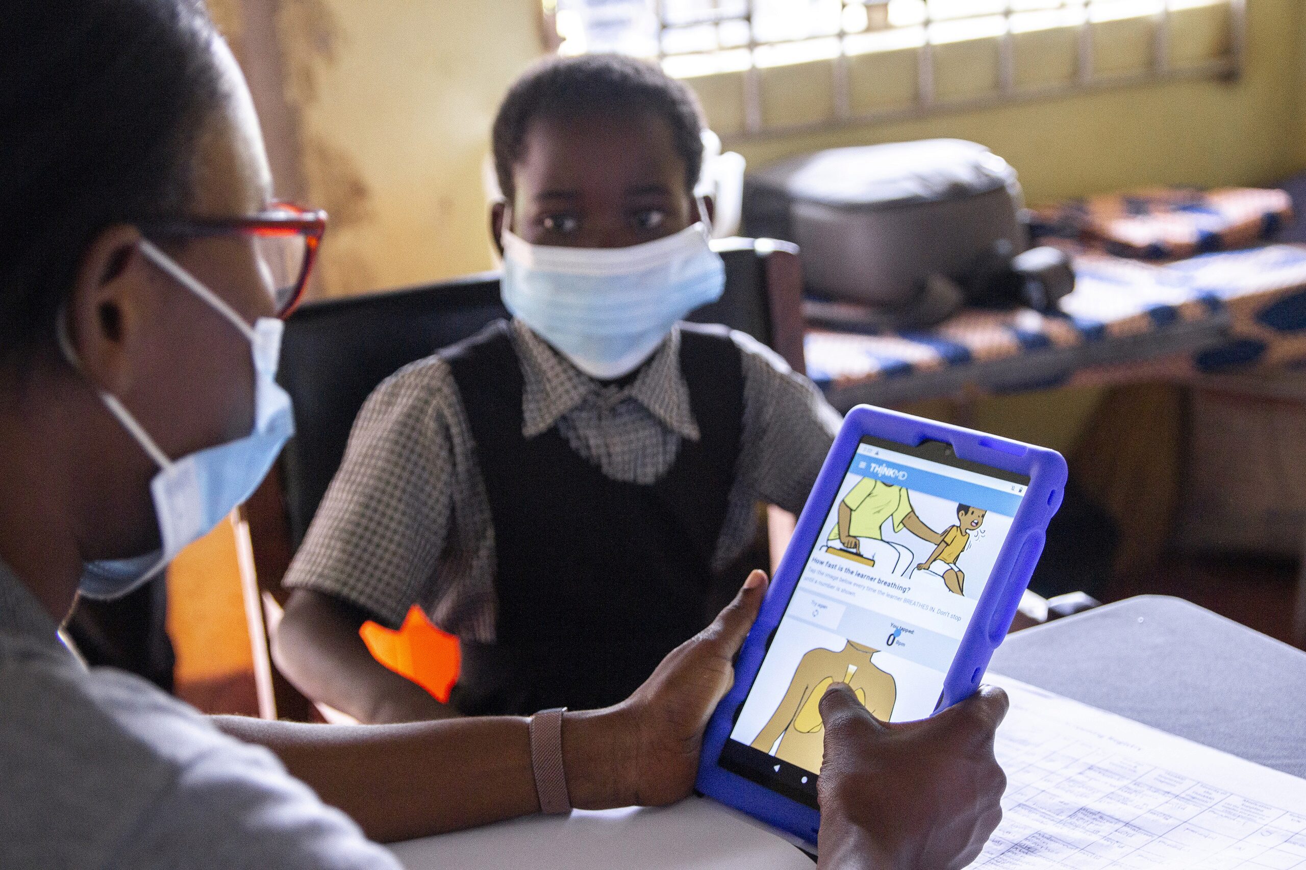 A person is showing a child an educational app on a tablet in a classroom setting. Both are wearing face masks.