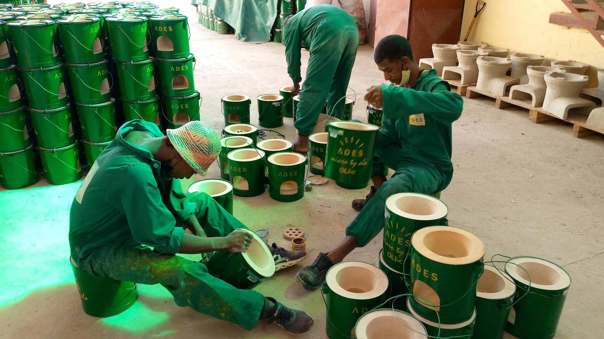 A few individuals in green uniforms assembling green ADES ceramic cookstoves in a workshop, with stacks of stoves in the background.