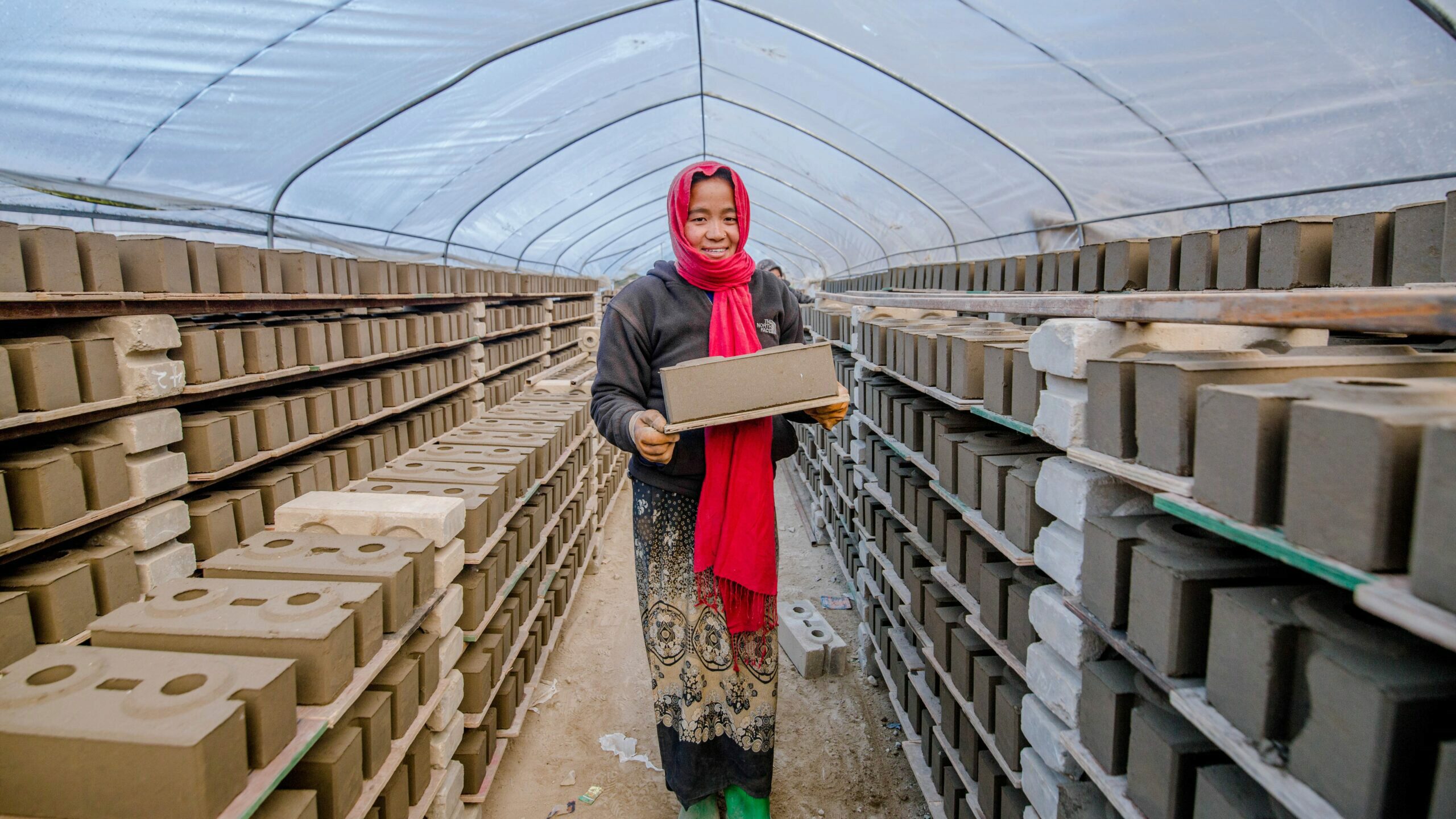 A person is standing in a tunnel-like structure filled with stacks of eco-friendly bricks, carrying one brick and smiling.