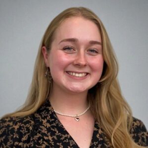 A headshot of a smiling person with long, blonde hair wearing a patterned top and a pearl necklace.