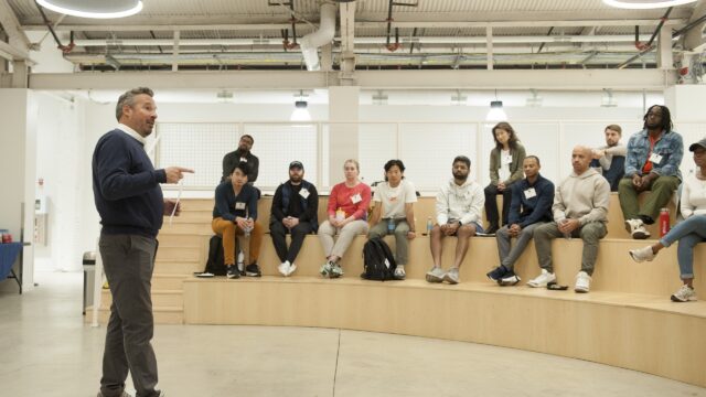 A person is speaking to a group sitting on tiered wooden seats in a modern interior space with industrial lighting and exposed pipes.