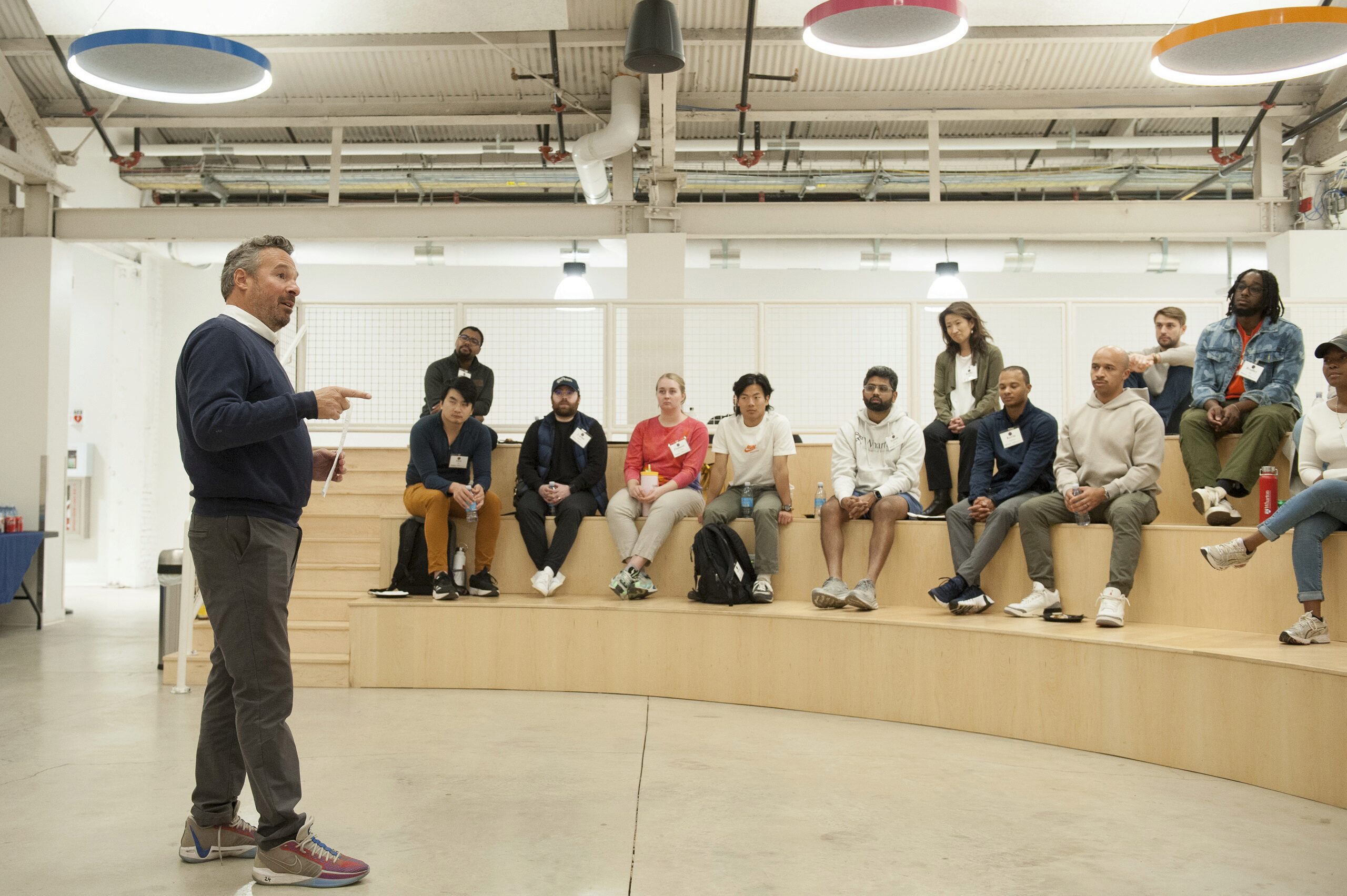 A person is speaking to a group sitting on tiered wooden seats in a modern interior space with industrial lighting and exposed pipes.