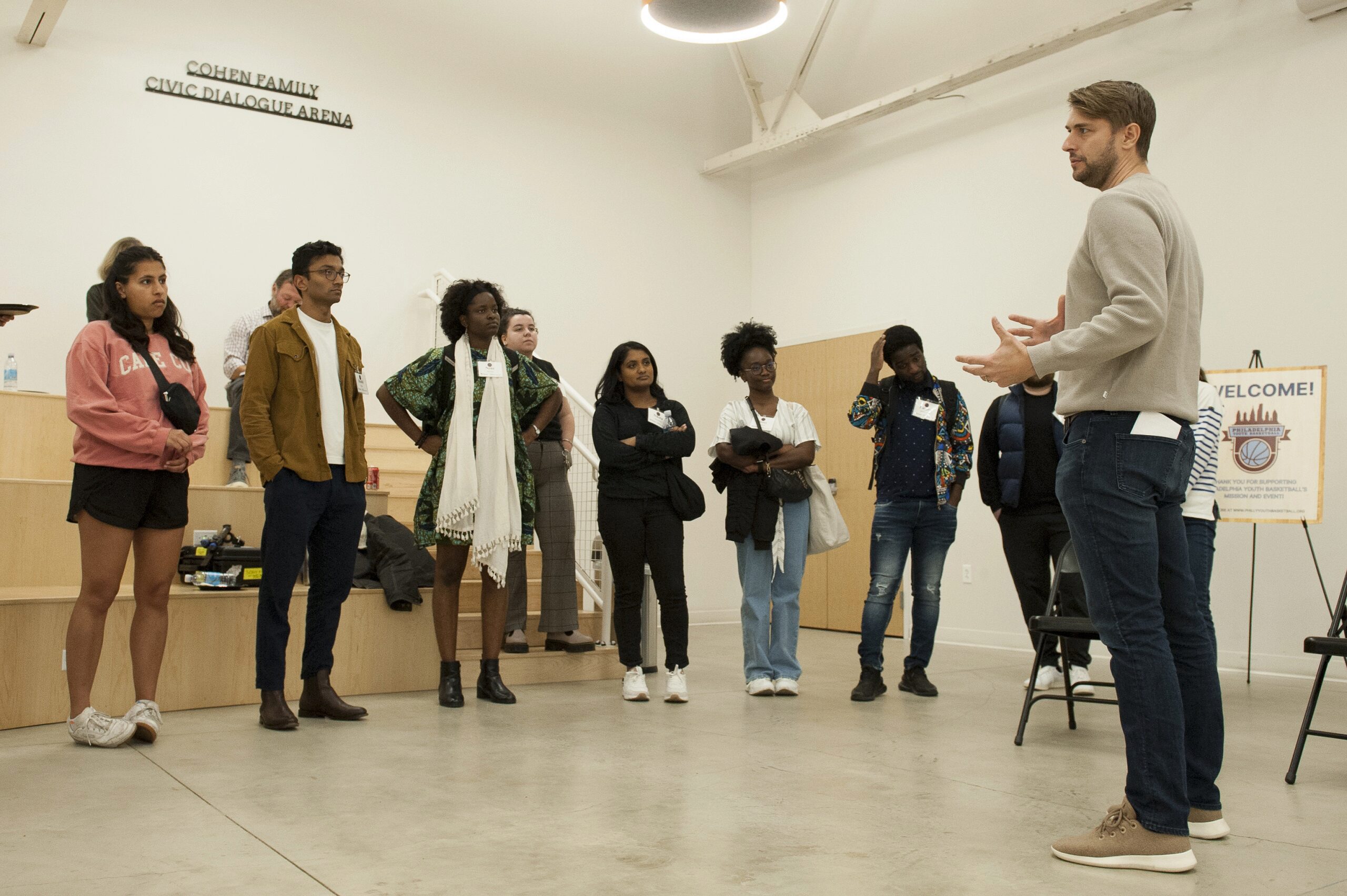 A speaker is addressing a diverse group of individuals standing in a room labeled "Cohen Family Civic Dialogue Arena." The atmosphere suggests a workshop or discussion event.