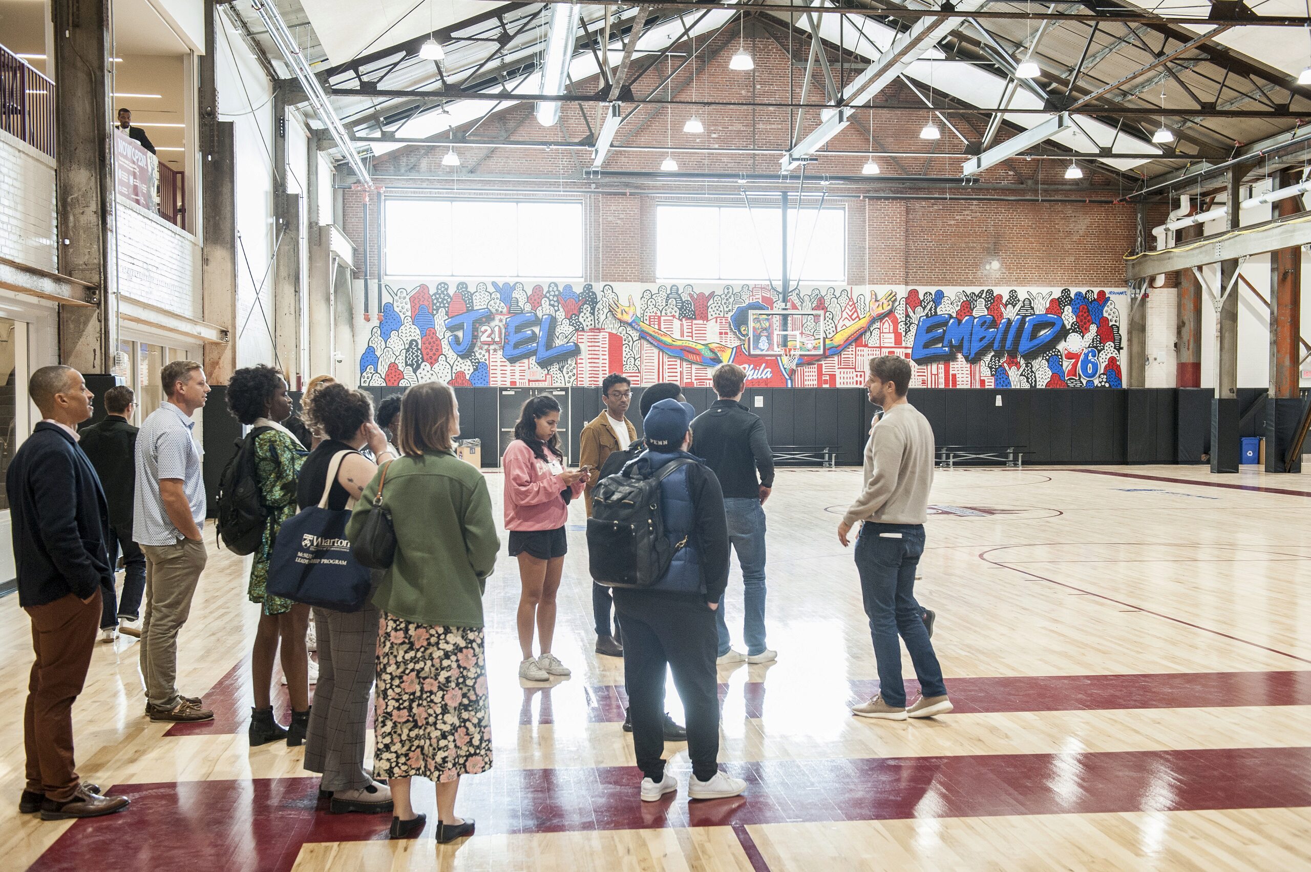 A group of people stands in an indoor basketball court with a hardwood floor. The wall features colorful graffiti art with the name "Embiid" and the number 21, alongside urban imagery. The setting appears to be modern and well-lit.