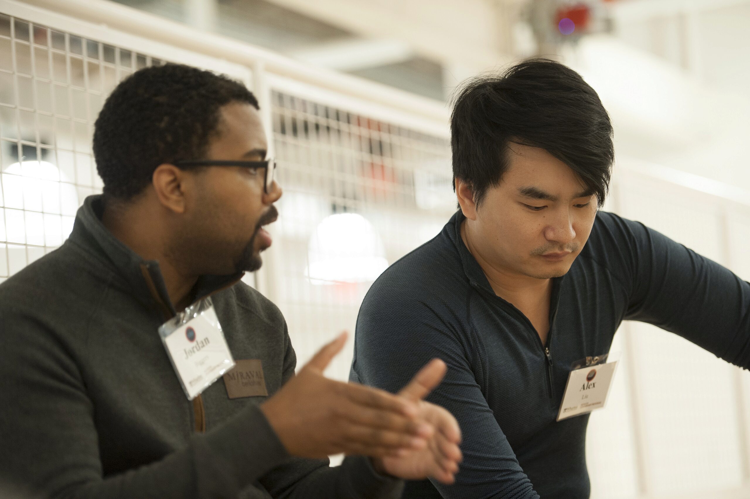 A candid moment between two people in conversation, possibly in a professional or educational setting. One individual is speaking, gesturing with their hands, while the other listens attentively. Both wear name badges.