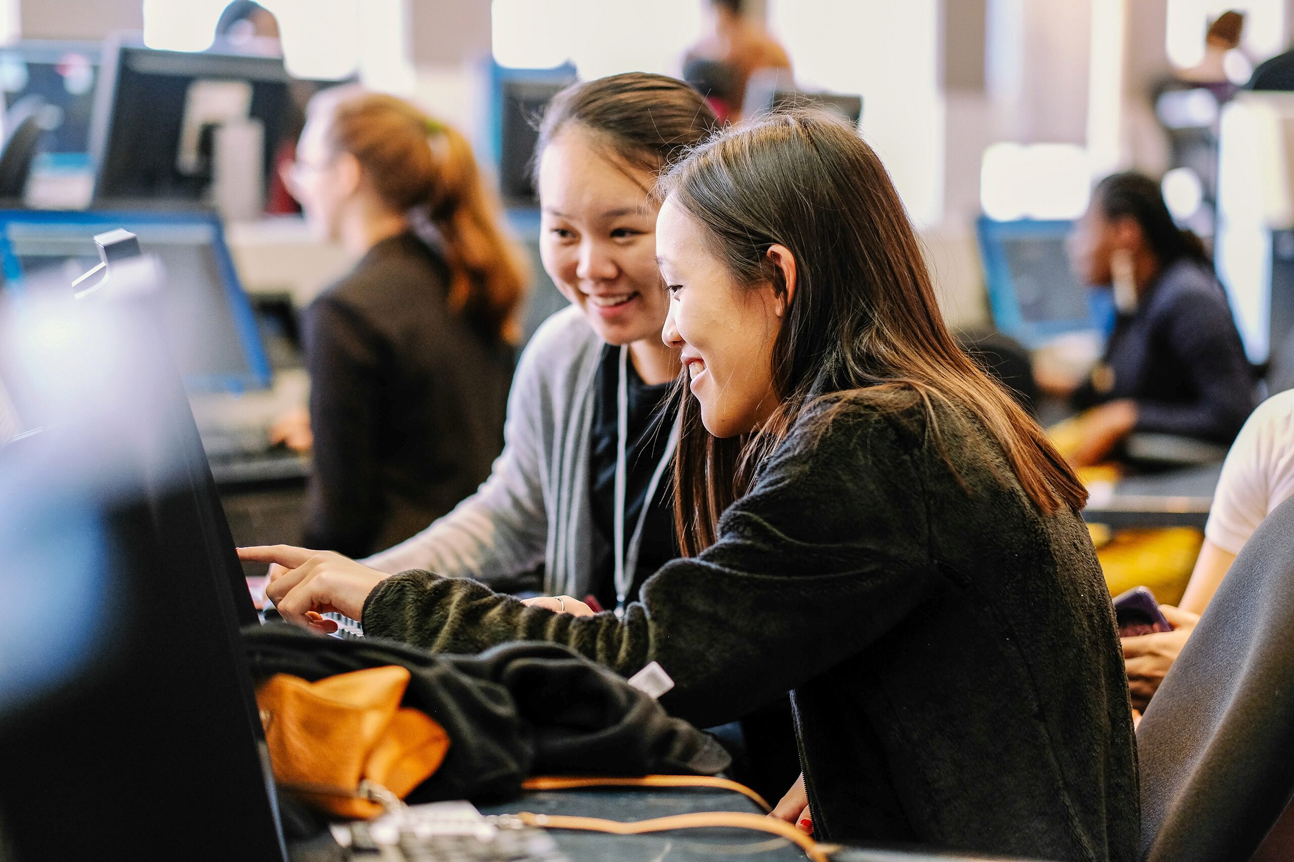 A couple of people are smiling and engaging with a computer in an office or classroom setting. Several others are working at computers in the background.