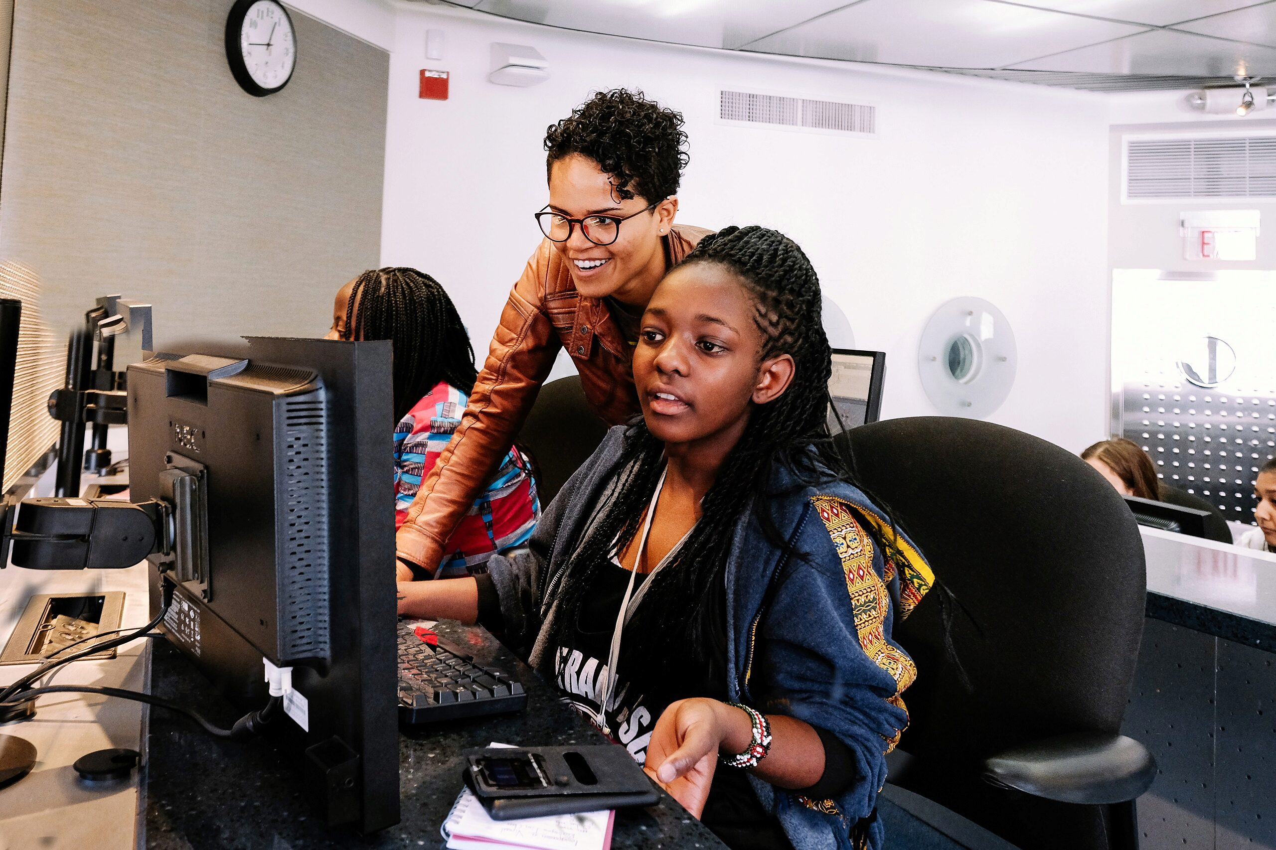 A person stands over another, who is seated at a computer in an office or learning environment. They're both focused on the screen, and appear to be collaborating or learning. Several other people are in the background.