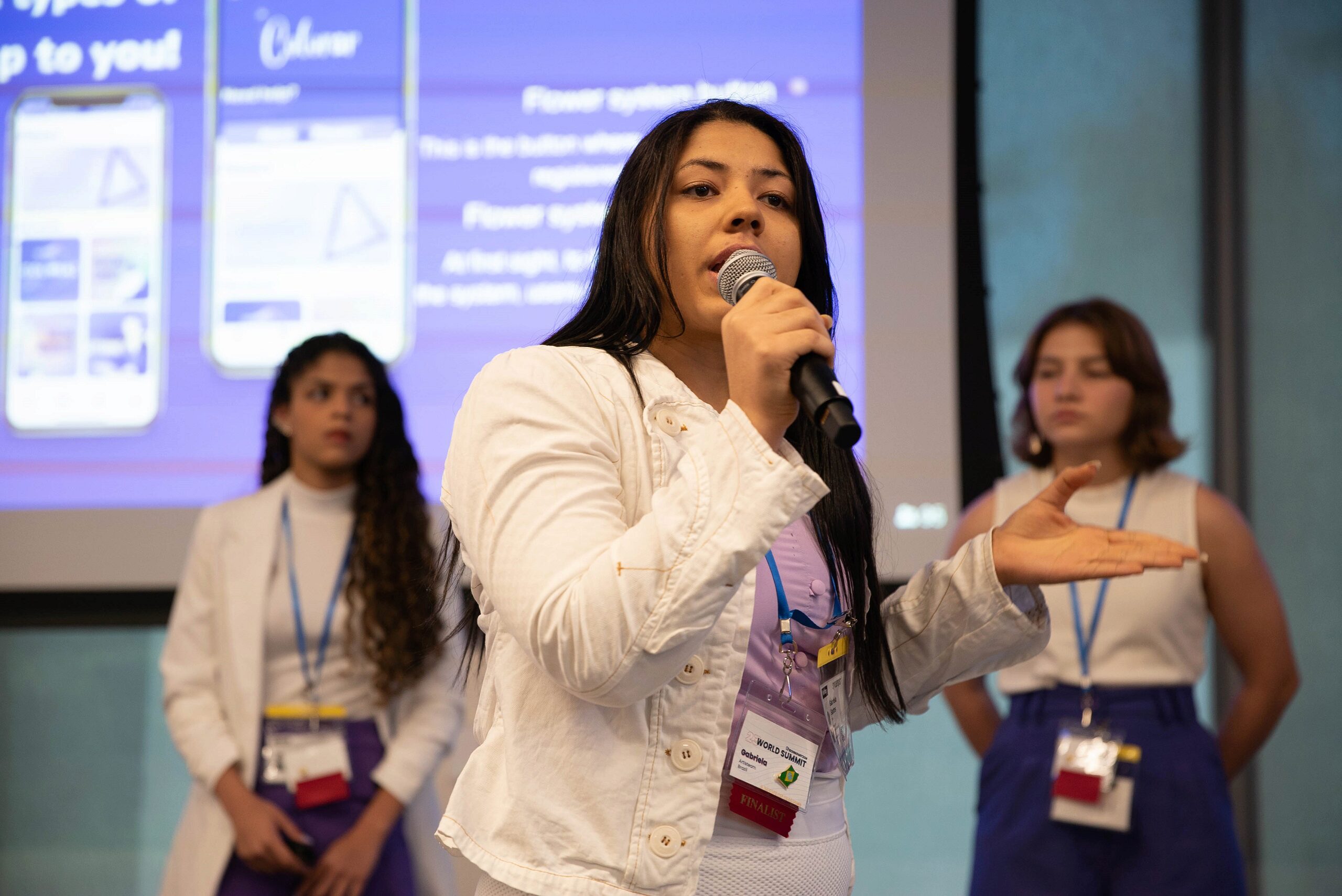 A person holding a microphone is speaking in front of a presentation screen, with two others standing in the background. All are wearing name tags, suggesting a professional or academic event.