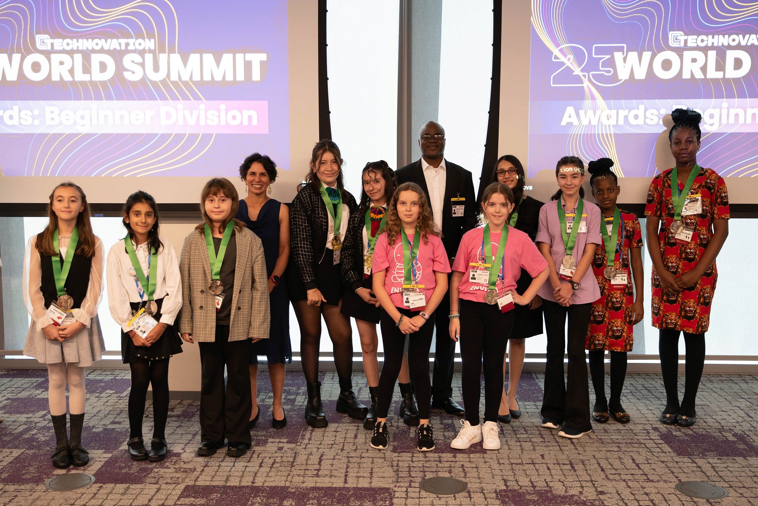 A group of people posing together, with some wearing medals. They are in front of displays that read "Technovation World Summit" and "Awards: Beginner Division." The setting suggests an award ceremony or conference event.