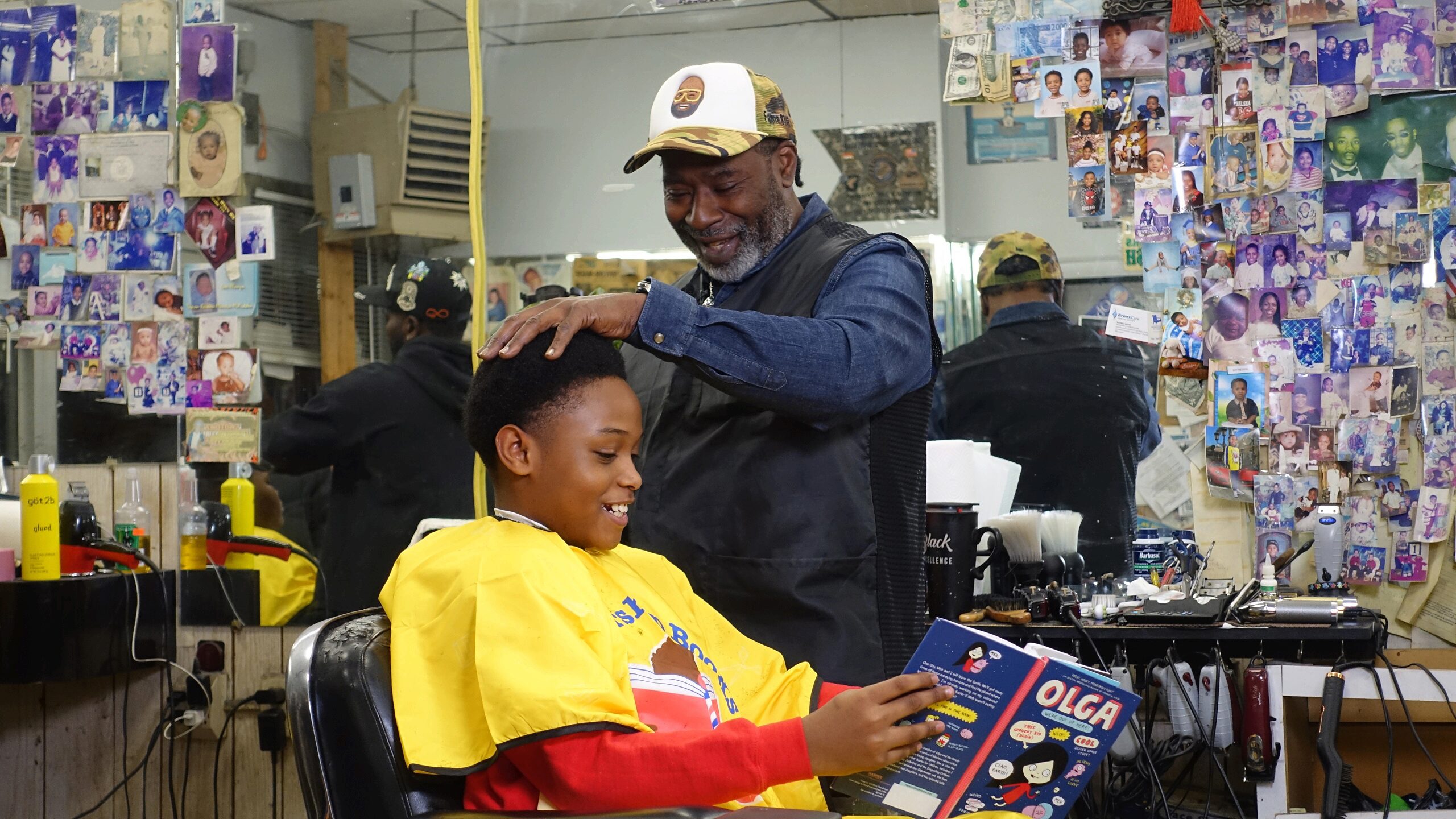 A barber is trimming a smiling child's hair while the child reads a book. The barber shop is decorated with numerous photos on the wall.