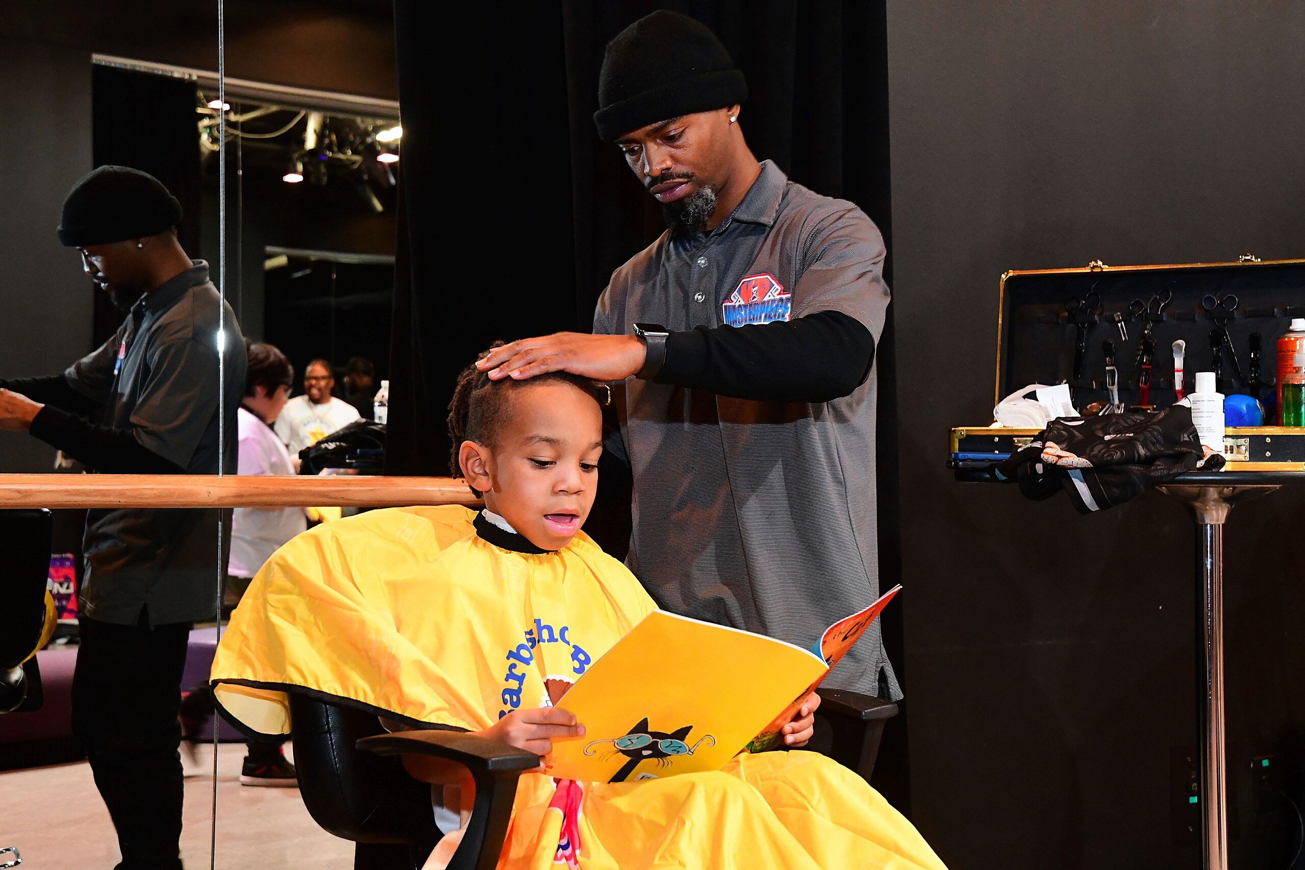 A child in a yellow barber cape reads a book while sitting in a barber chair. A barber places a hand on the child's head, with barber tools visible nearby.