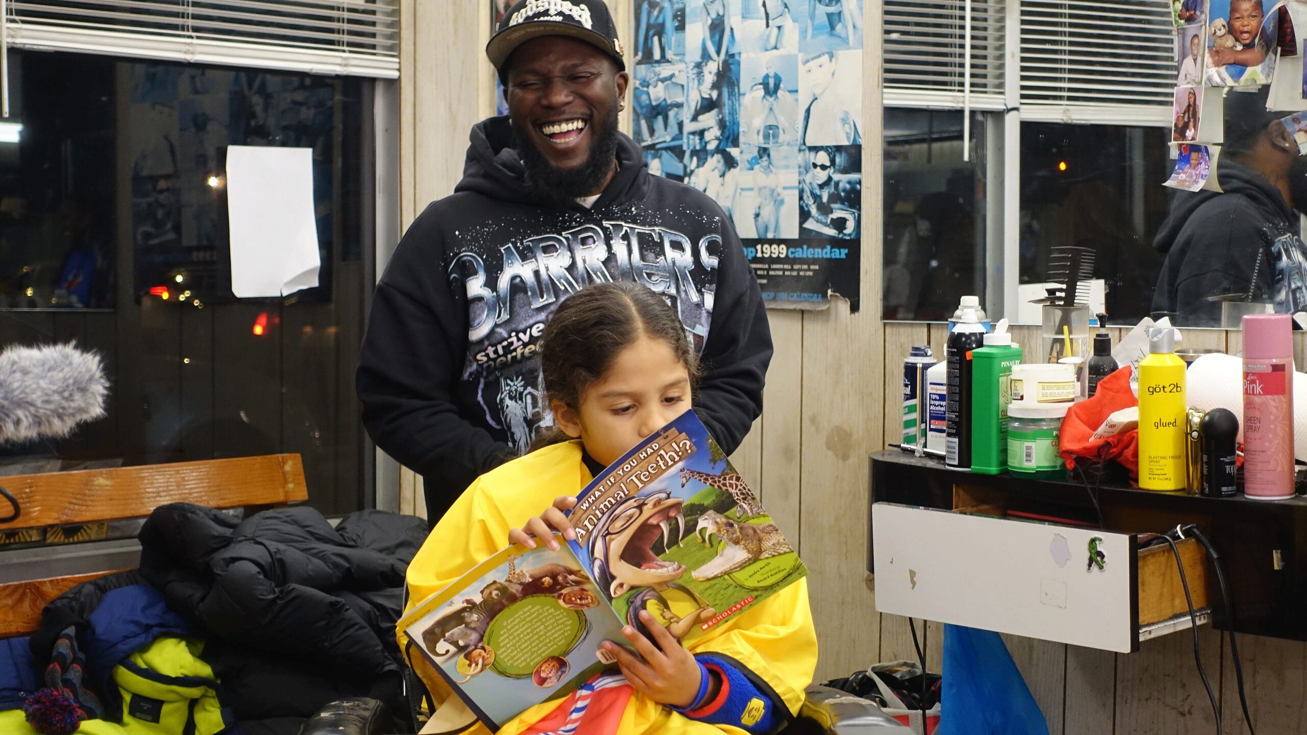 A person is cutting another person's hair in a barbershop. The person in the chair is reading a children's book titled "What If You Had Animal Teeth?" The atmosphere seems relaxed and joyful, with colorful products visible on the counter.