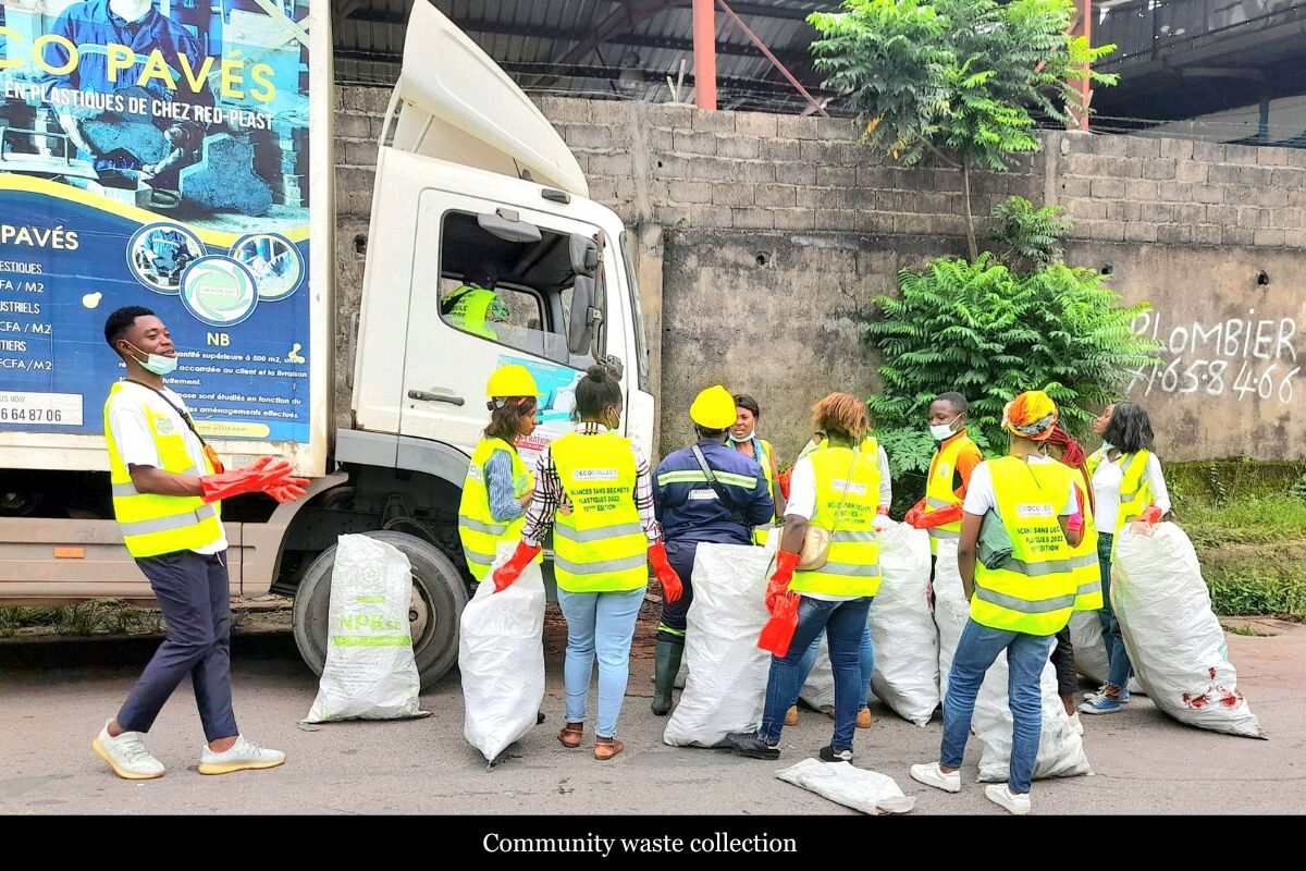A group of people wearing yellow safety vests and helmets, holding bags and standing near a truck, engaged in community waste collection.