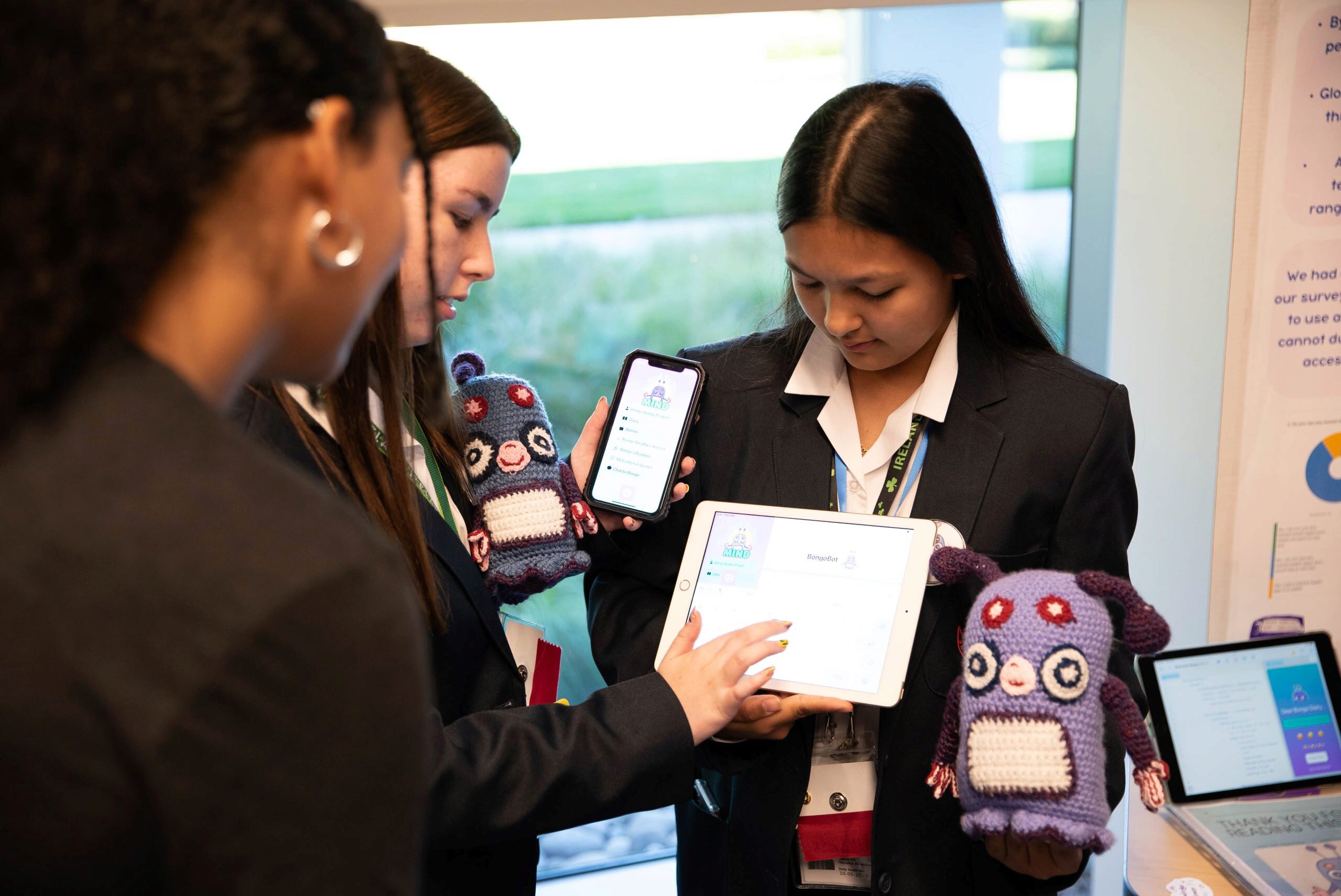 A small group of people in business attire is examining a tablet and smartphone displaying a digital application. They also hold plush toys resembling robots or creatures.