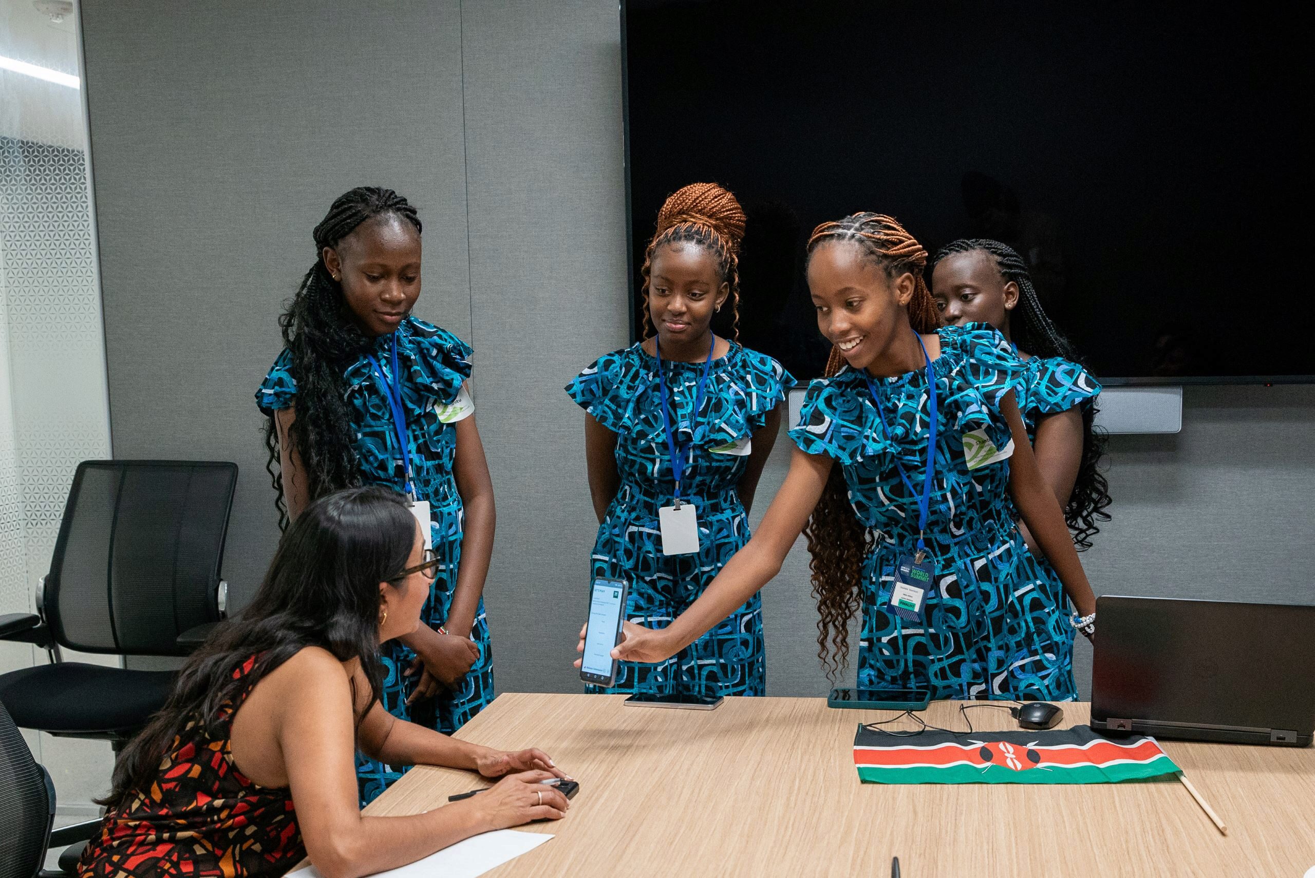 A group of people in patterned blue outfits stand around a table, showing a smartphone to a seated person. A Kenyan flag and a laptop are on the table.