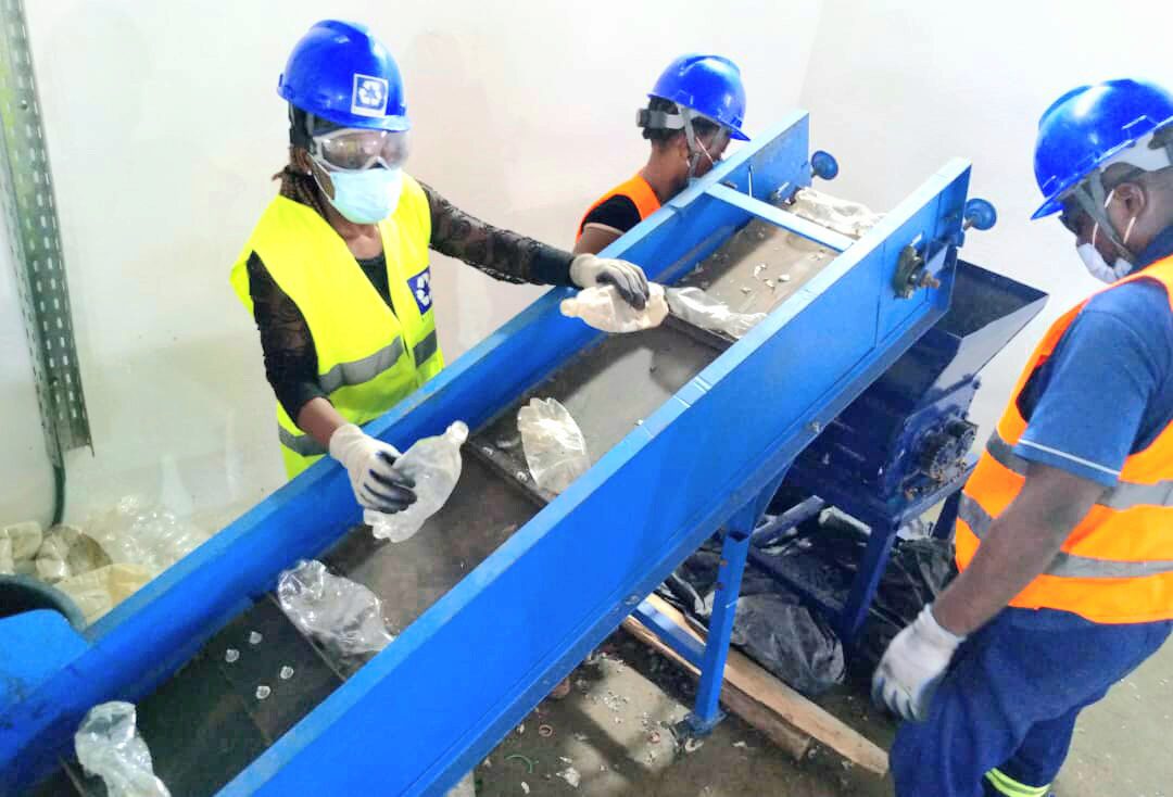 A small group of people wearing safety gear is sorting plastic bottles at a recycling facility. They are using a conveyor belt system.