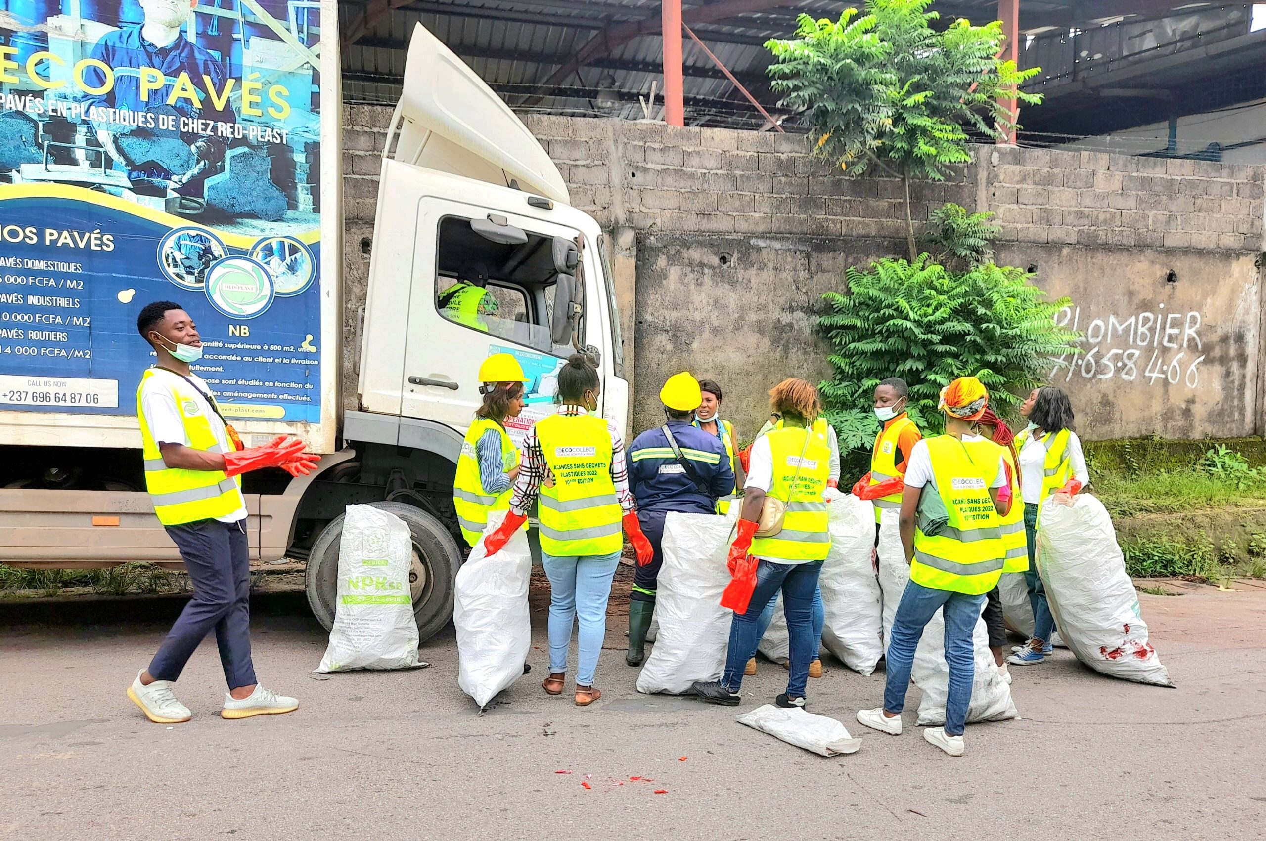 A group of people wearing yellow safety vests and hard hats stand near a truck, holding large sacks. They appear to be involved in a recycling or cleaning activity.