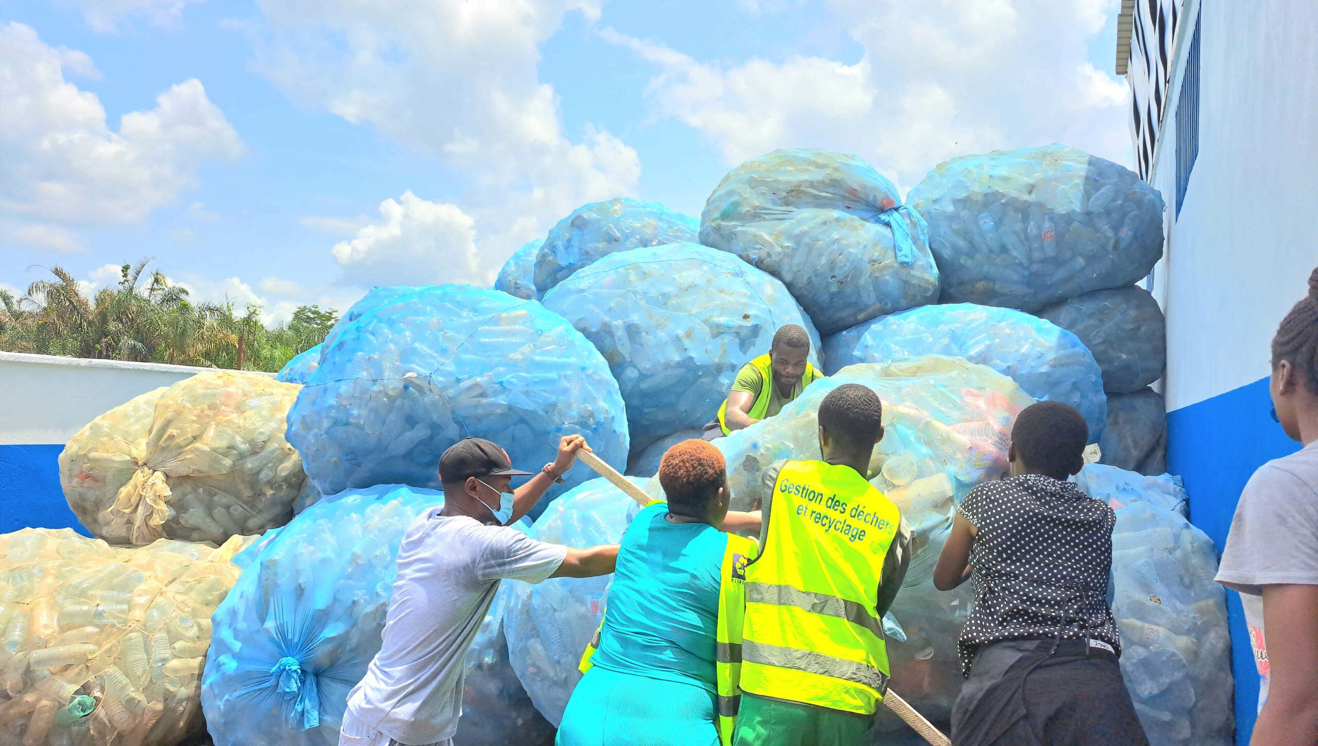 A group of people is handling large bags filled with plastic bottles, likely for recycling purposes. The bags are stacked in an outdoor setting.