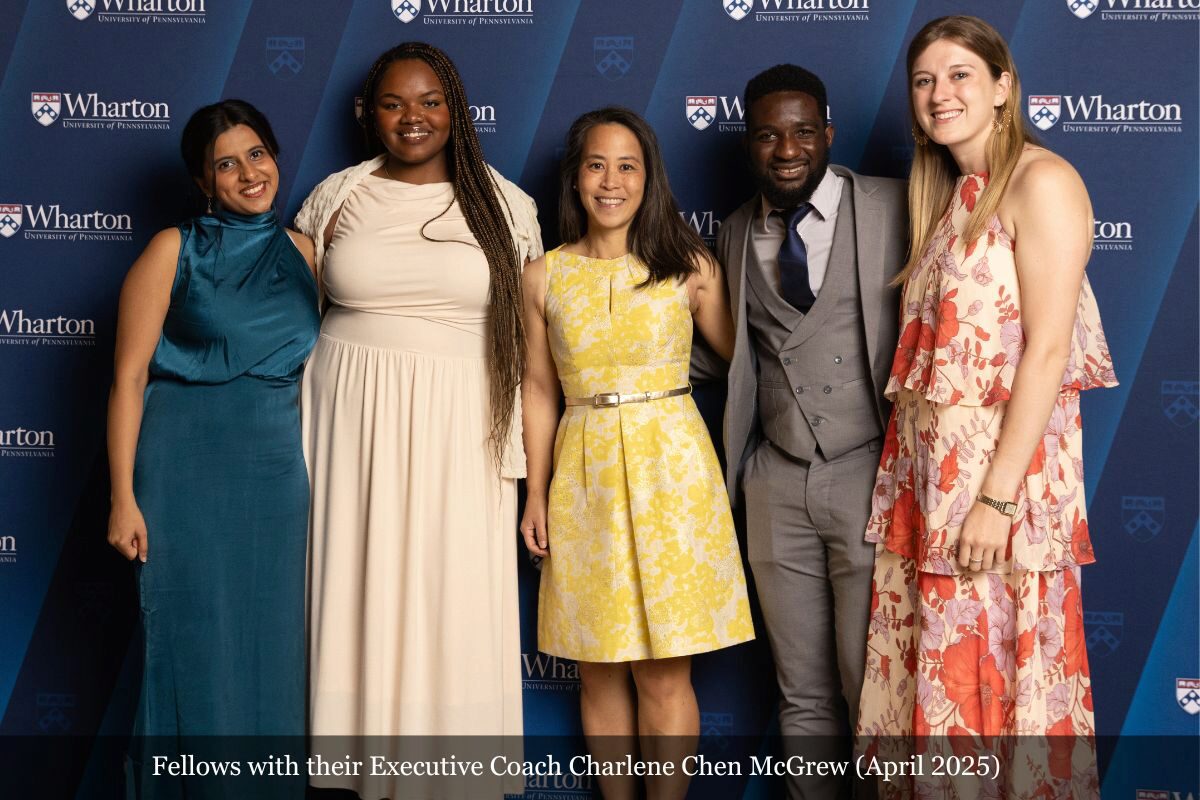 A group of five individuals smiling in front of a backdrop with the Wharton School logo. They are dressed in formal attire, suggesting a professional or celebratory event.