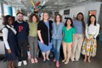 A group of people smiling and posing together in an office environment. Various colorful decorations and workstations are visible in the background.