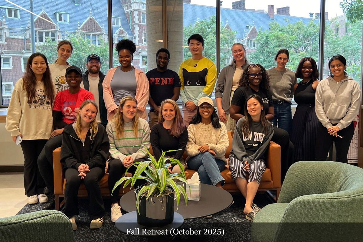 A diverse group of people smiling and posing indoors for a photo, with a large window showing a building in the background. Labeled "Fall Retreat (October 2025)."