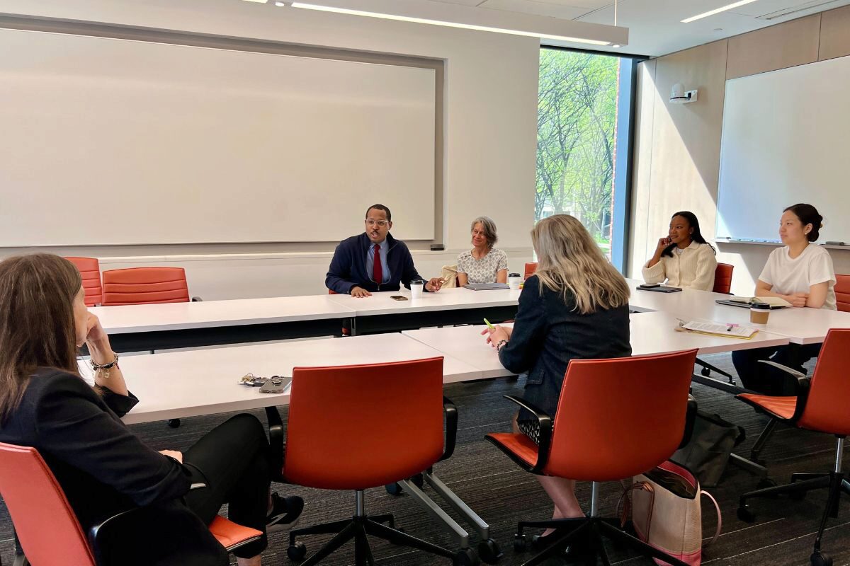 2 A group of people are engaged in a meeting in a conference room with red chairs around a white table. A large window and whiteboard are visible.