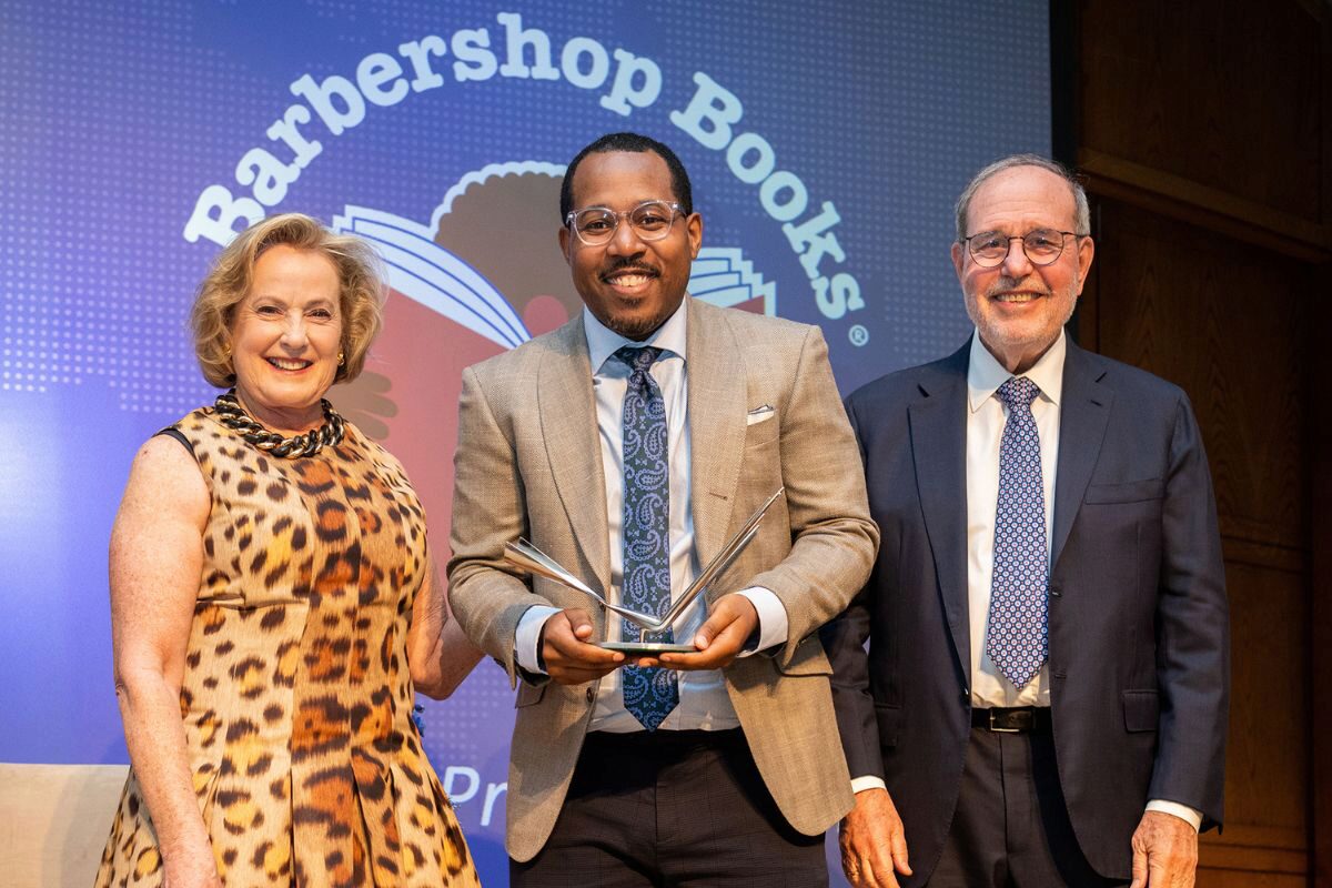 2025 Resized Lipman Winner Images (1) A small group of people posing in front of a Barbershop Books logo. The person in the center is holding a trophy and is flanked by two others, all smiling.