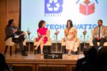 A panel discussion with four individuals sitting on stage, each holding a microphone. The backdrop features logos and the word "TECHNOVATION".