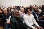 A diverse audience seated in a conference or seminar setting, with attendees engaged and listening attentively. Some hold event materials or name badges.