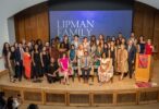 A large group of people posing on a stage with a banner in the background reading "Lipman Family Prize." Several individuals are seated in front, with others standing around them. A podium with a Wharton School banner is visible to the right.