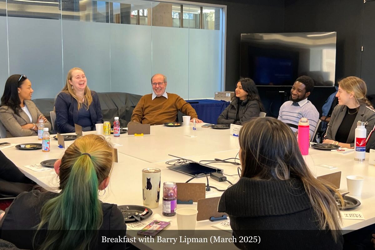 A small group of people seated around a conference table, engaging in conversation and smiling. The setting appears to be a business or academic environment.